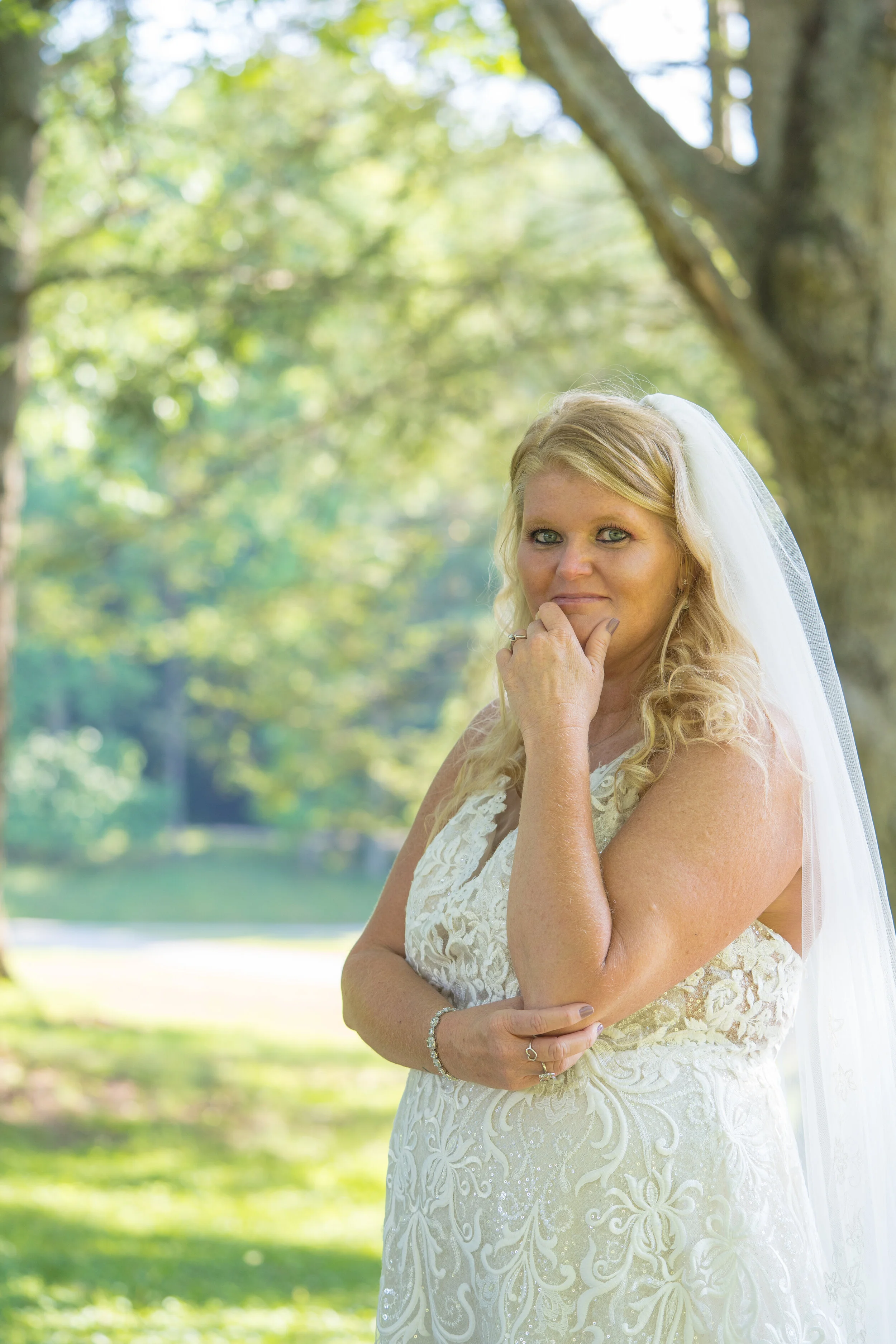 A bride with blonde curly hair wearing a white lace wedding dress and veil, standing outdoors with trees in the background.
