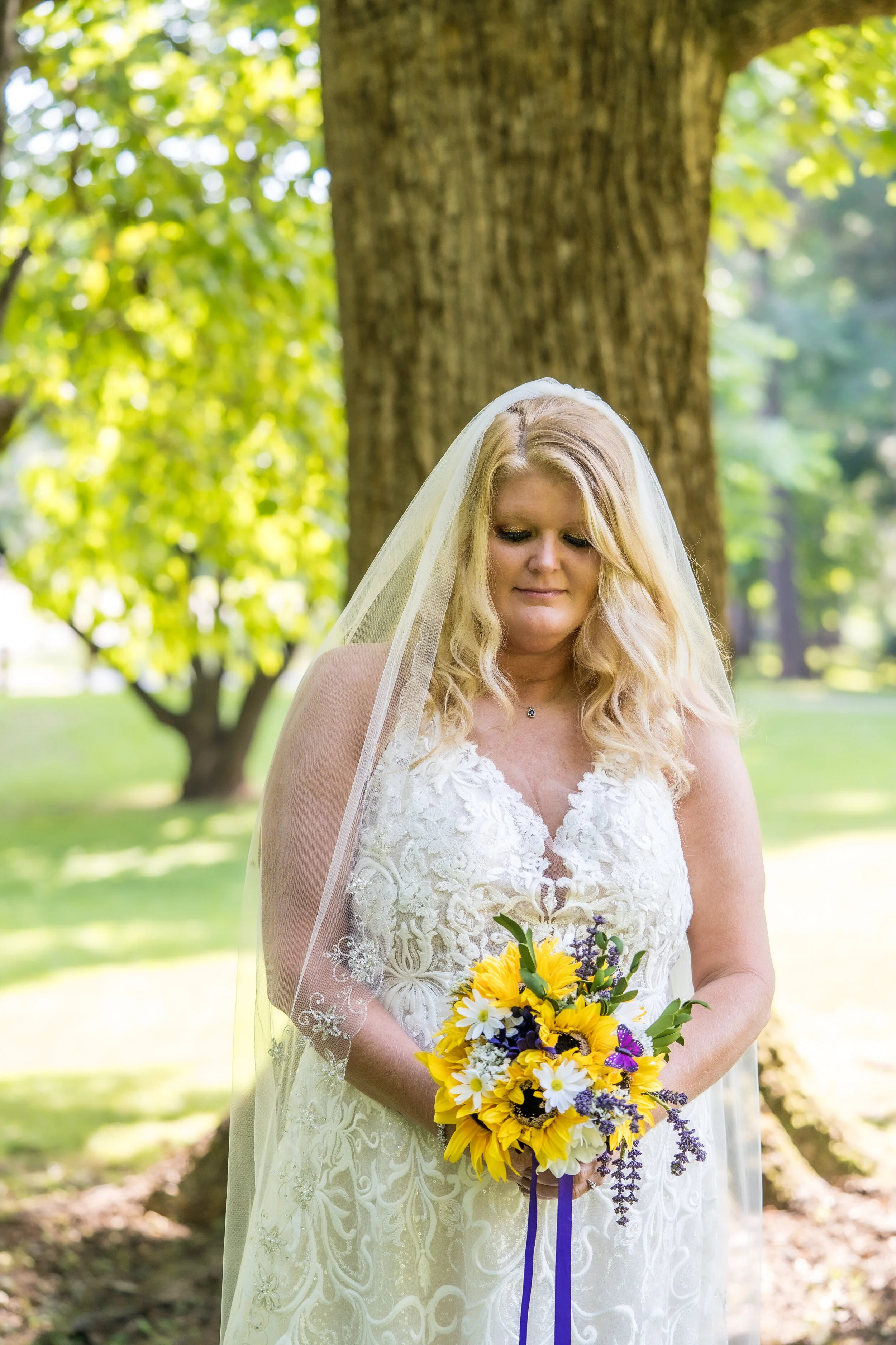 A bride in a white lace wedding dress and veil holds a bouquet of sunflowers, daisies, lavender, and purple accents, standing outdoors under a large tree with green leaves.