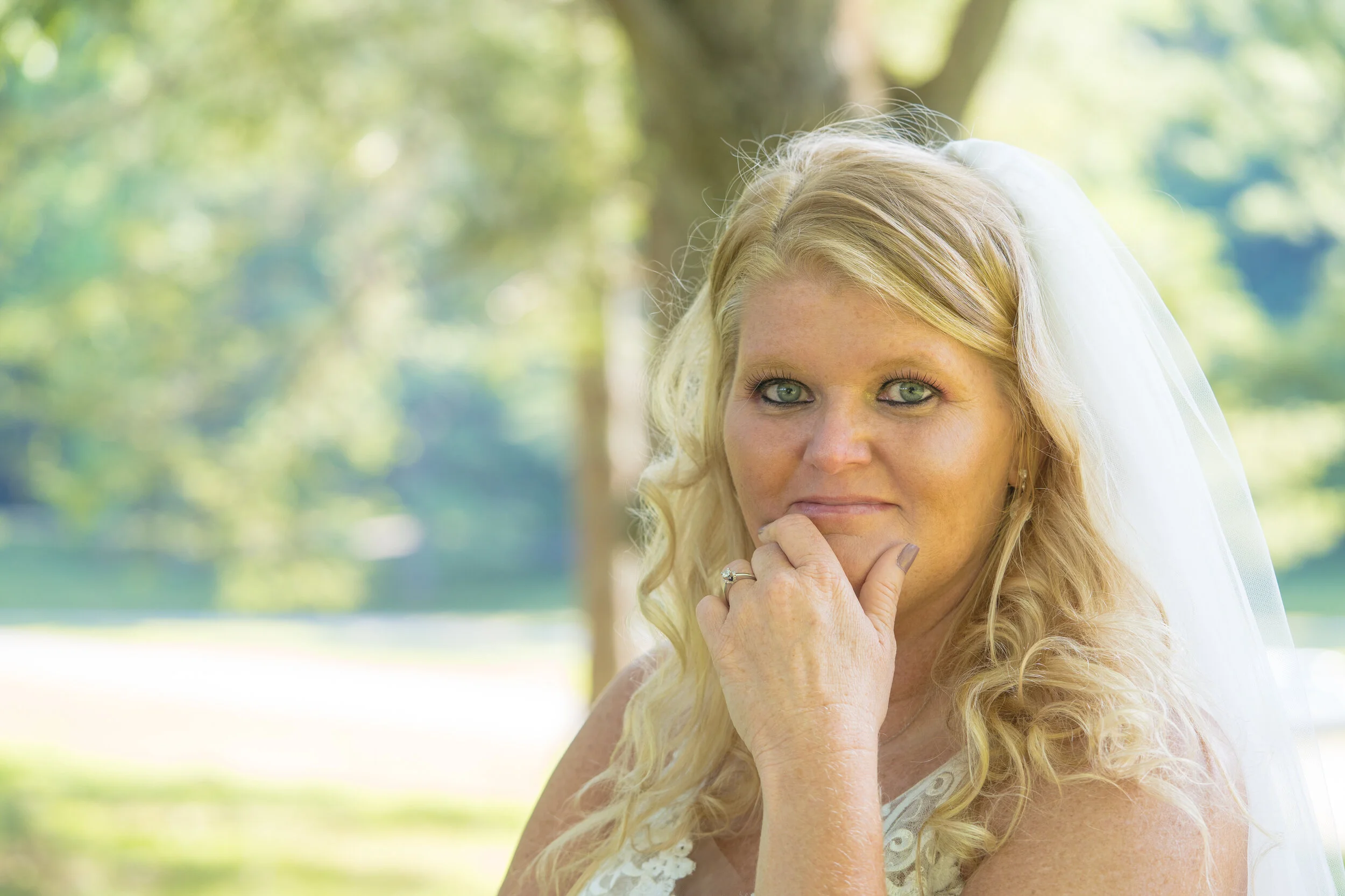 Close-up of a woman with blonde curly hair and blue eyes, wearing a wedding veil and white dress outdoors with greenery background.