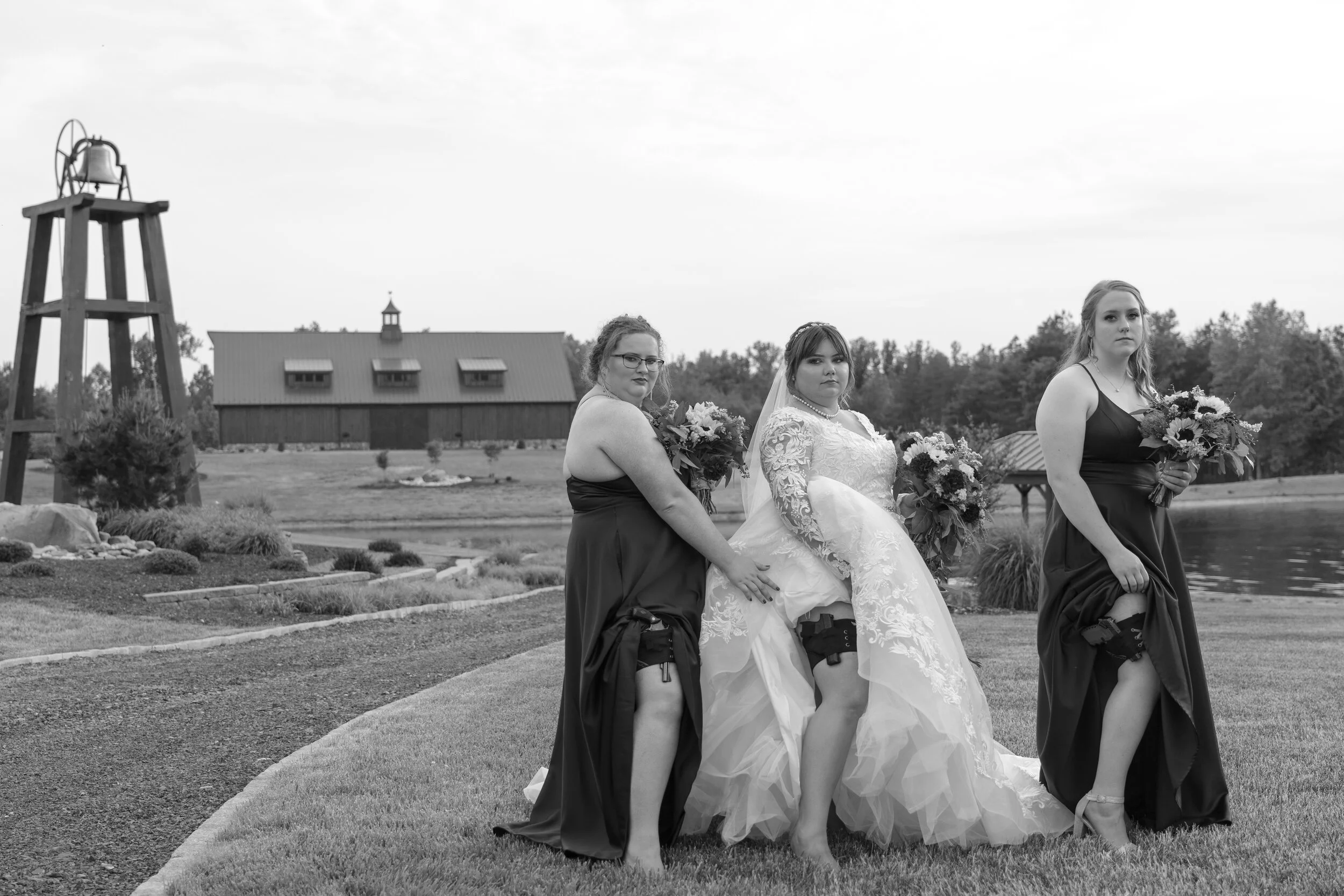 A bride in a wedding gown sitting on a bench with two bridesmaids, all holding bouquets, outdoors near a pond with a barn and water tower in the background, black and white photo.