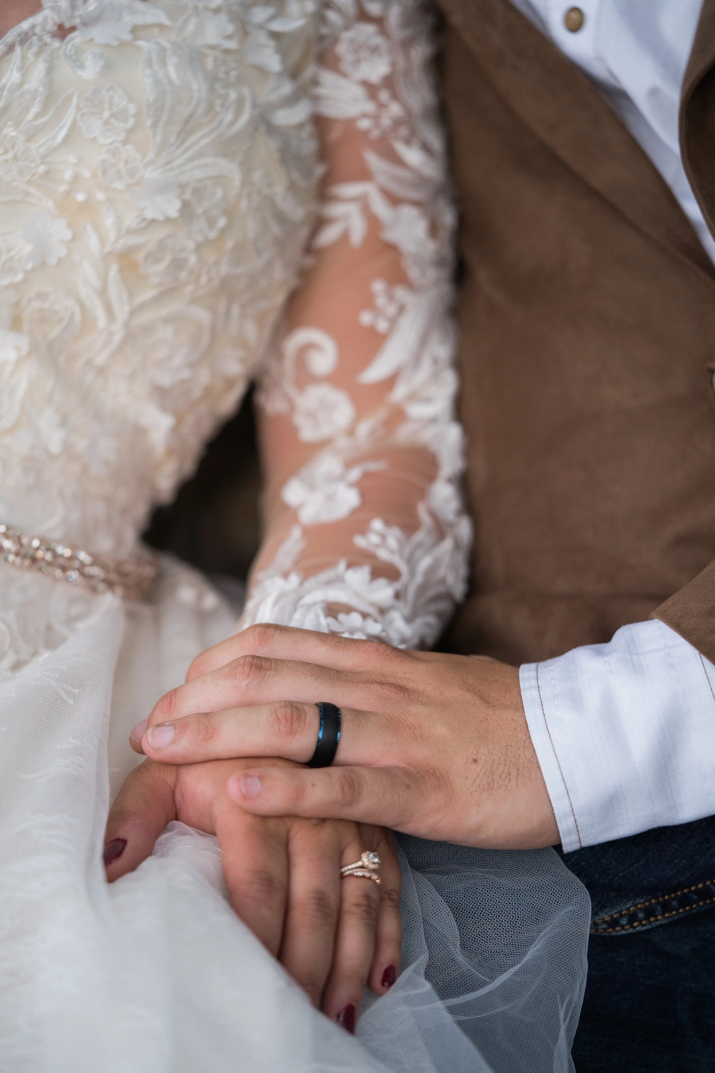 Close-up of a bride and groom holding hands during a wedding, with the bride wearing a lace wedding dress and rings on her fingers, and the groom wearing a white shirt, brown vest, and a black ring.