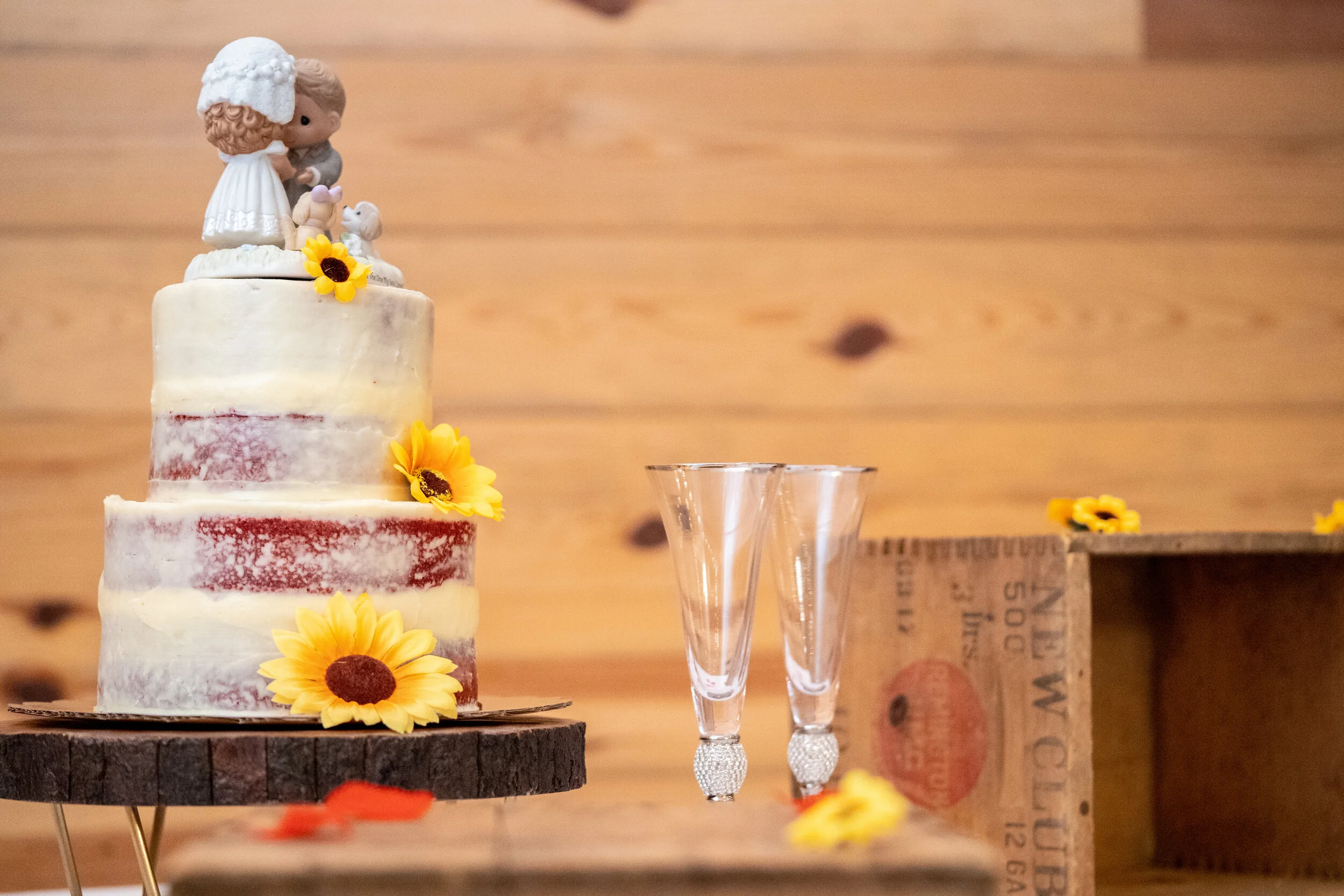 A three-tier wedding cake decorated with yellow sunflower flowers and a figurine topper of a girl holding a dog, set on a wooden stand, with two empty glass cups nearby on a rustic wooden table backdropped by a wood-paneled wall.