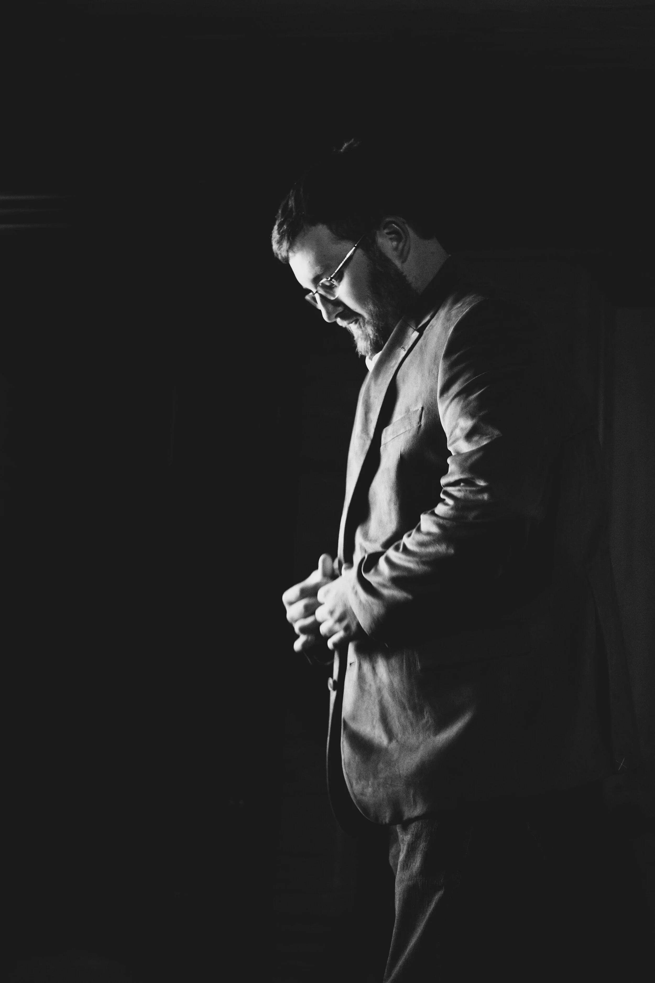 Black and white photo of a man in a suit adjusting his jacket, standing sideways with his head bowed.