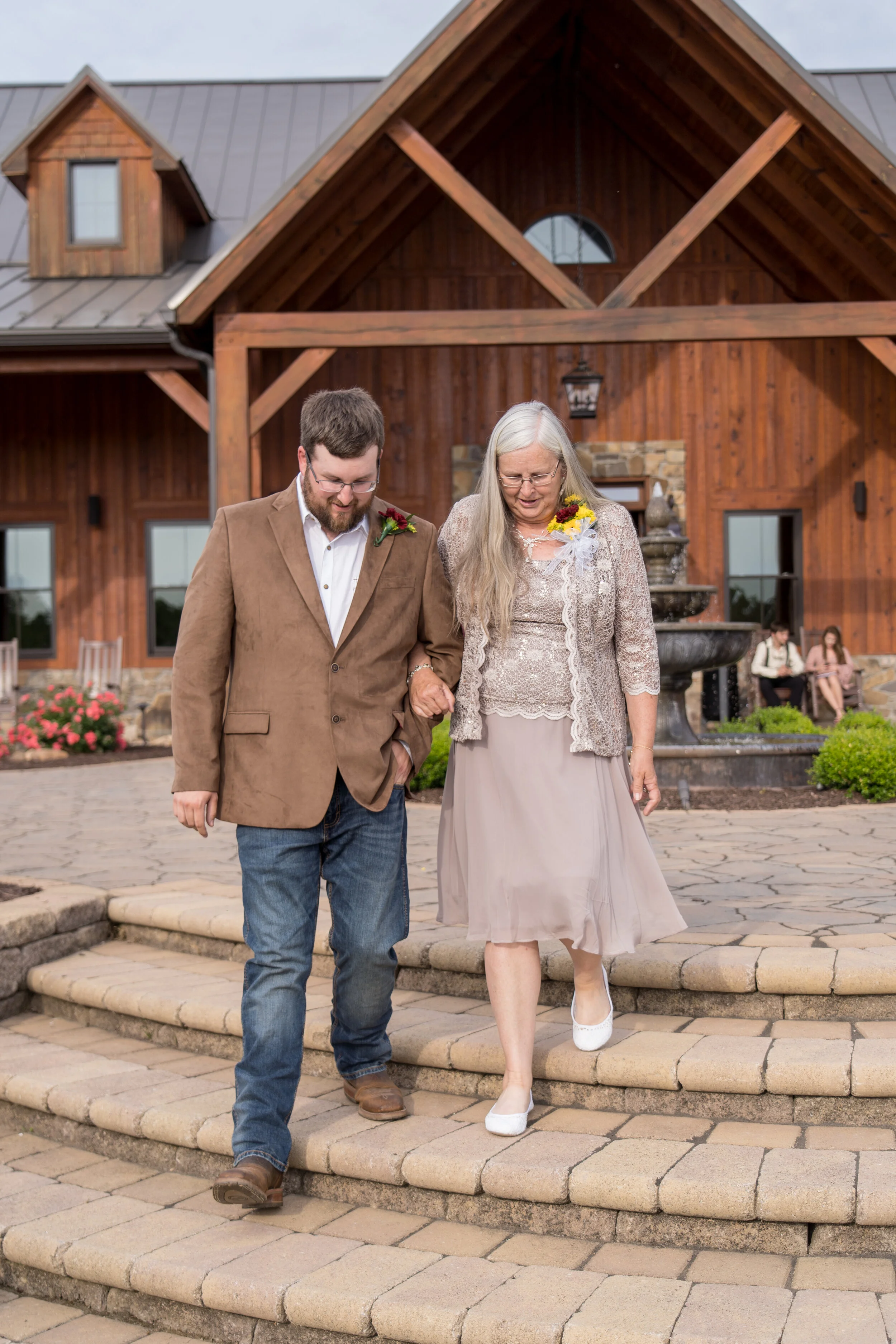 A young man and an older woman walking down steps outside a large wooden building, possibly at a wedding or special event. The man is wearing a brown blazer, white shirt, and jeans, while the woman is dressed in a light-colored dress with a lace top 