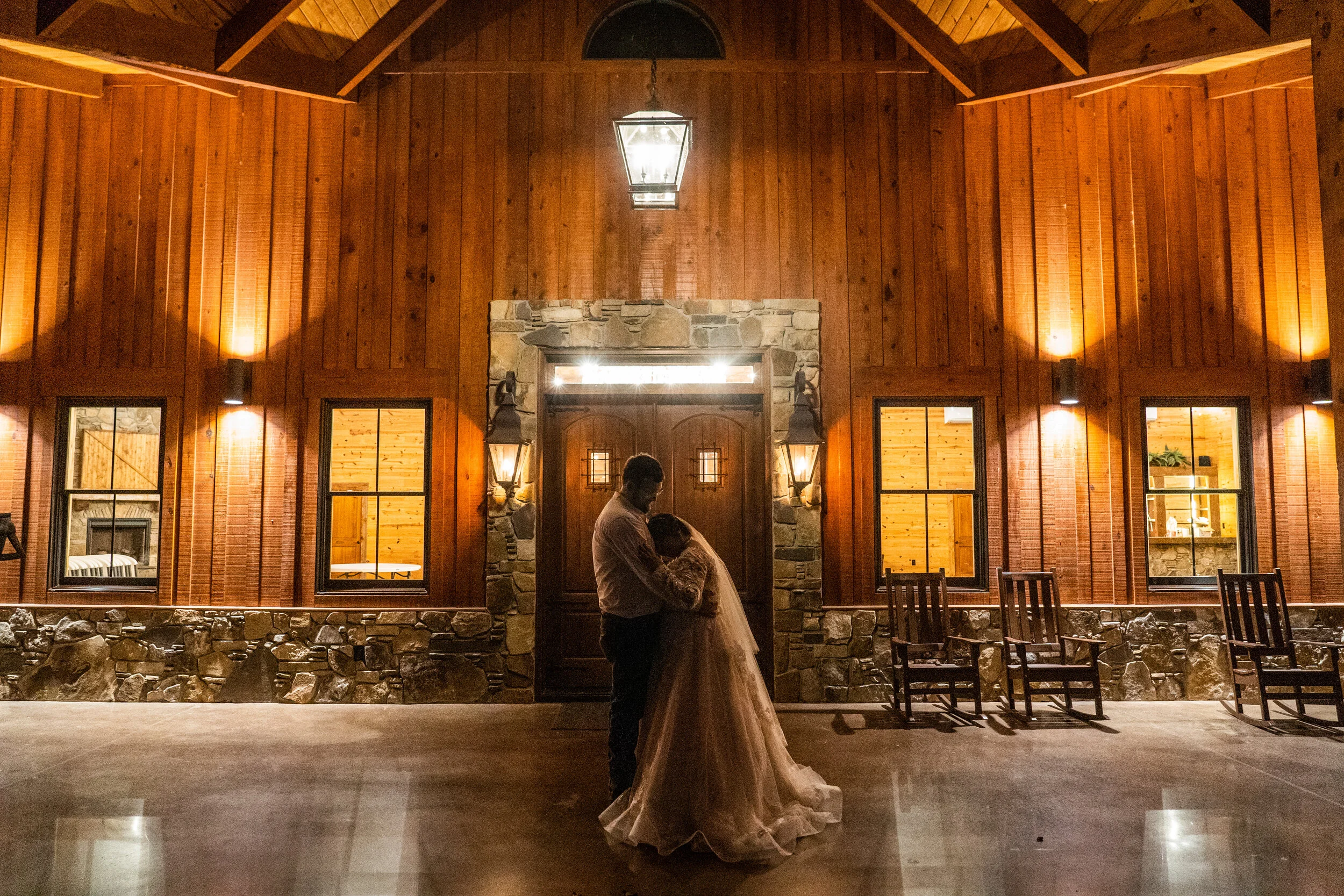 A couple dancing closely in front of a rustic wooden cabin outside at night, with warm lighting and chairs arranged along the sides.