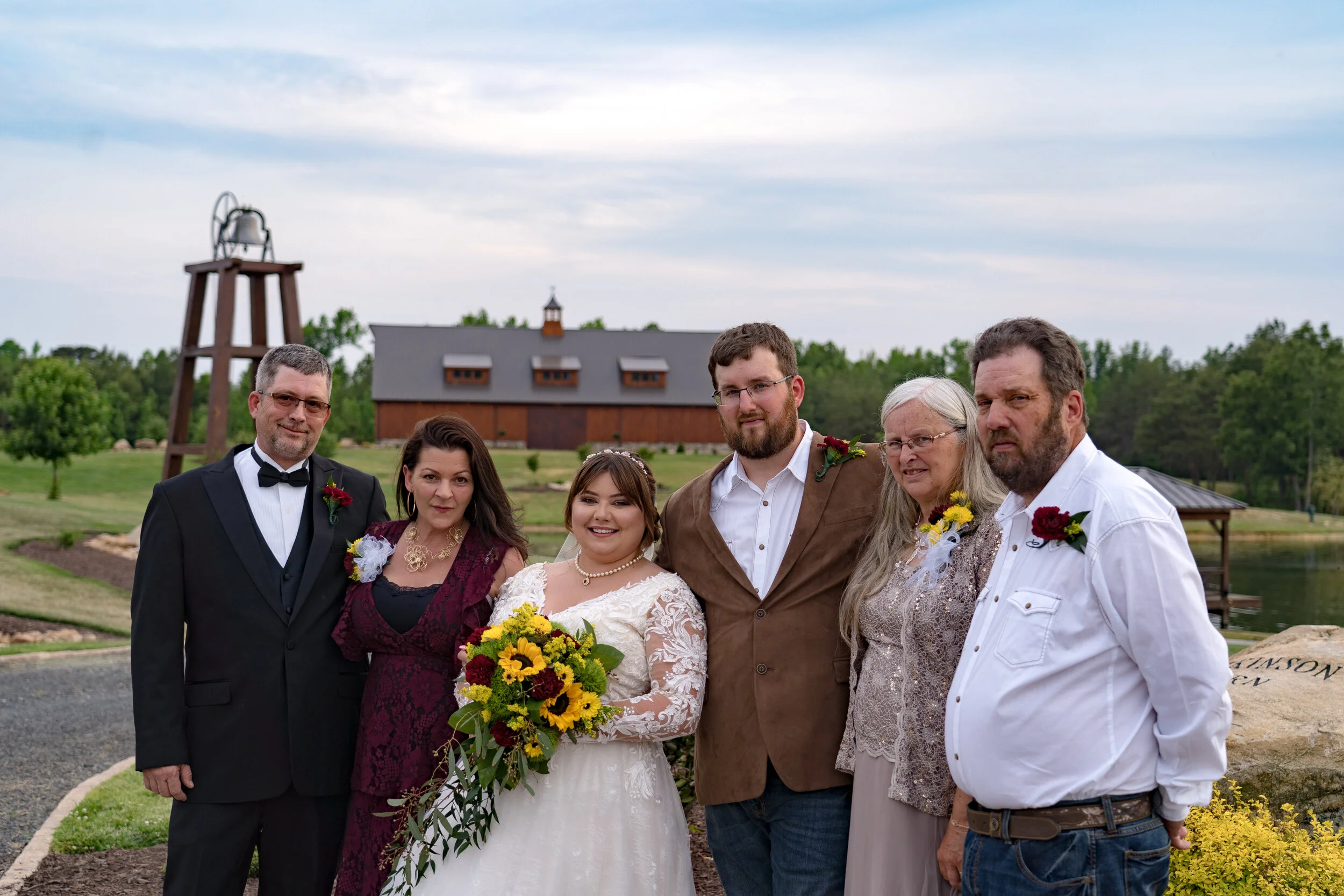 A group of six people, including a bride in a white dress holding a bouquet of yellow and red flowers, standing outdoors on a cloudy day with a rustic barn, trees, and a watermill in the background.
