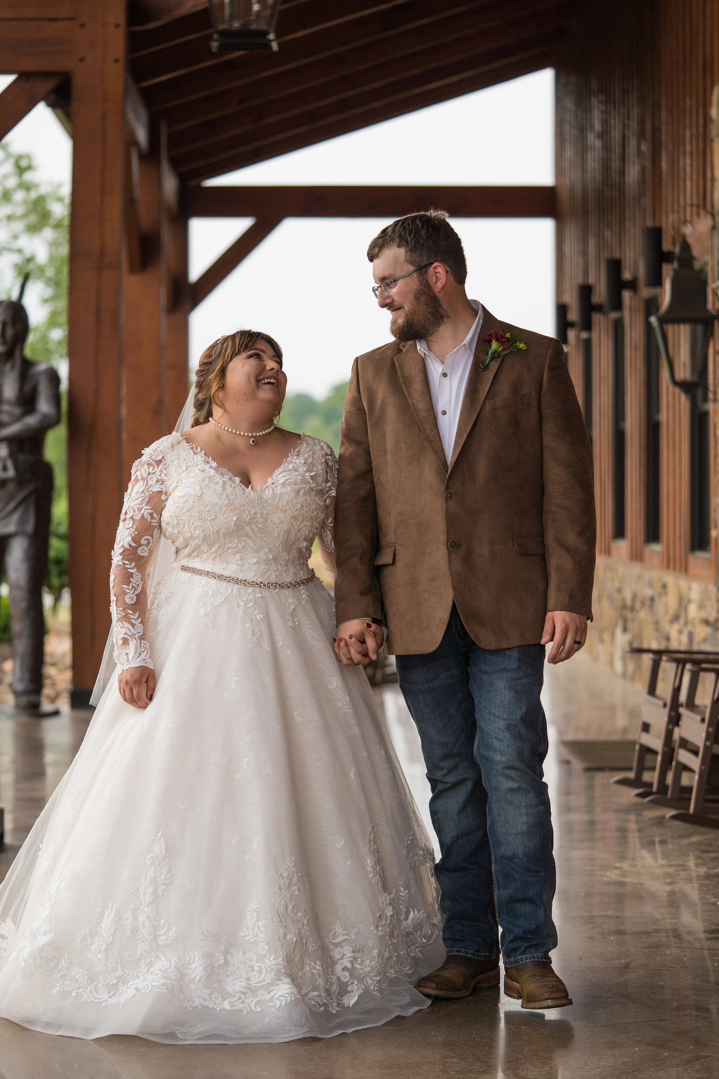 A bride and groom holding hands and smiling at each other during their wedding ceremony. The bride is wearing a white lace wedding gown with long sleeves and pearl jewelry. The groom is dressed in a brown blazer, white shirt, and jeans.