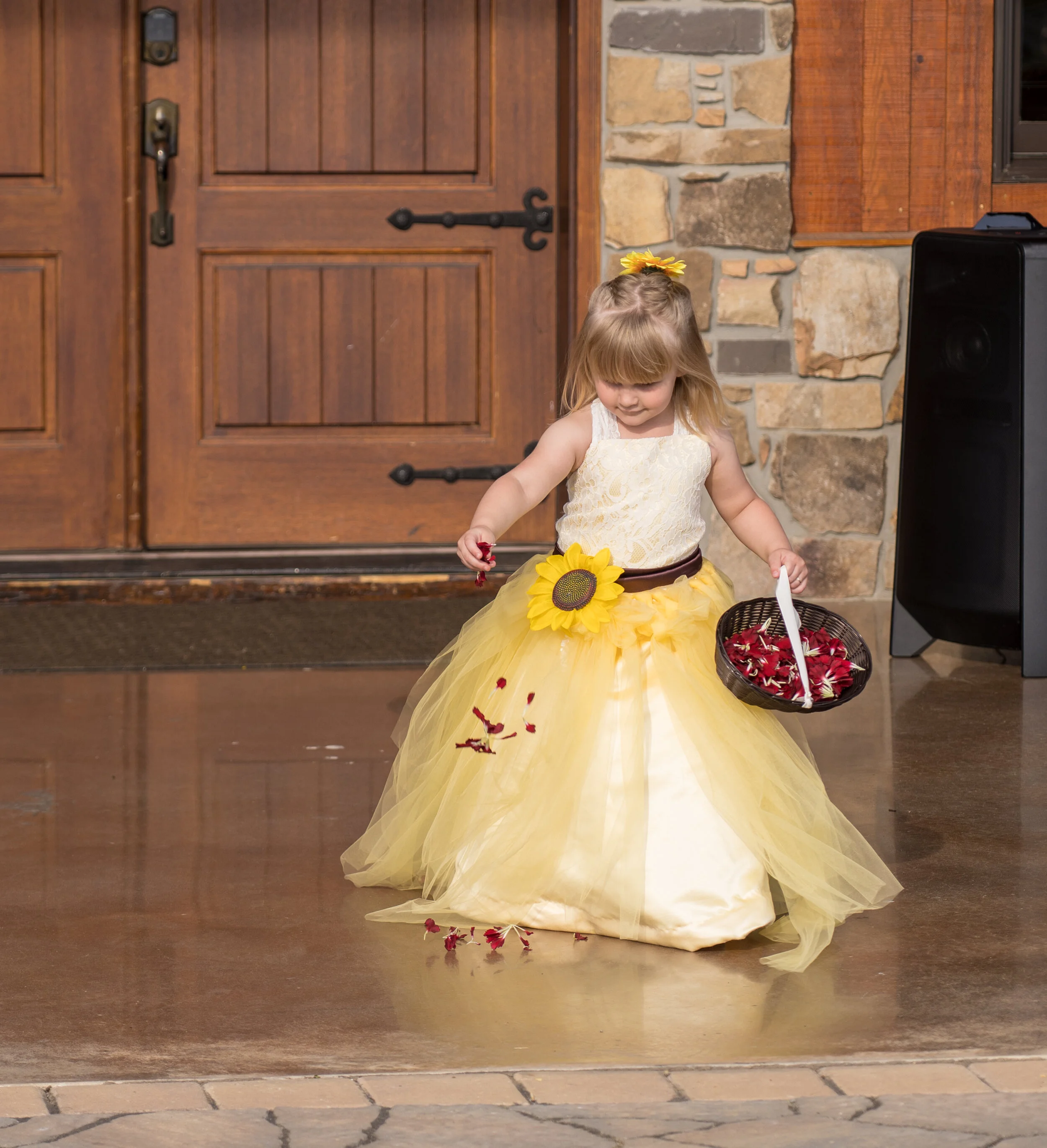 A young girl dressed in a yellow and white flower girl dress, holding a basket of red rose petals, standing in front of a wooden door with stone accents.
