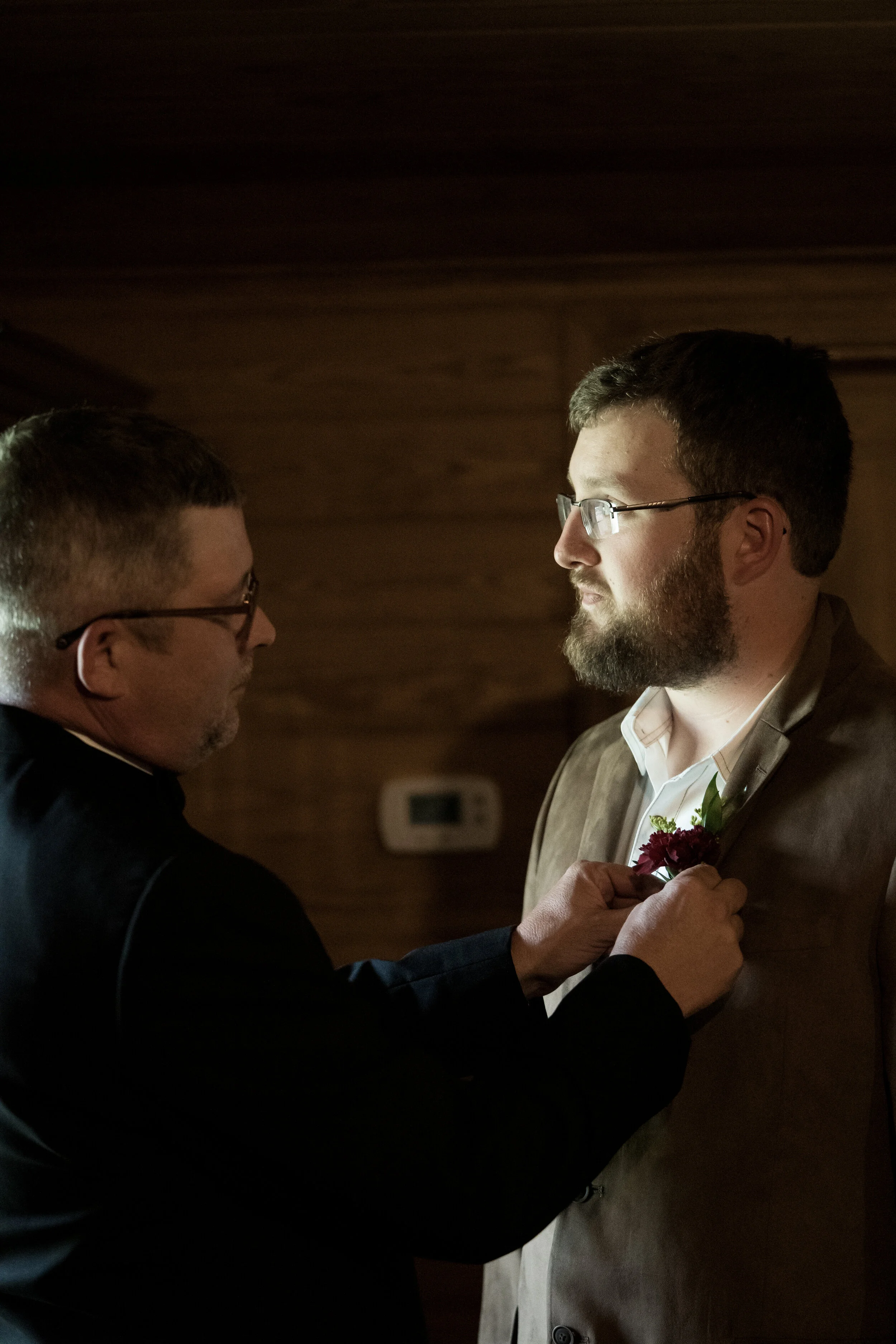 A man with a beard and glasses, wearing a suit jacket, is being pinned with a boutonniere by another man with glasses, in an indoor setting with wooden walls.