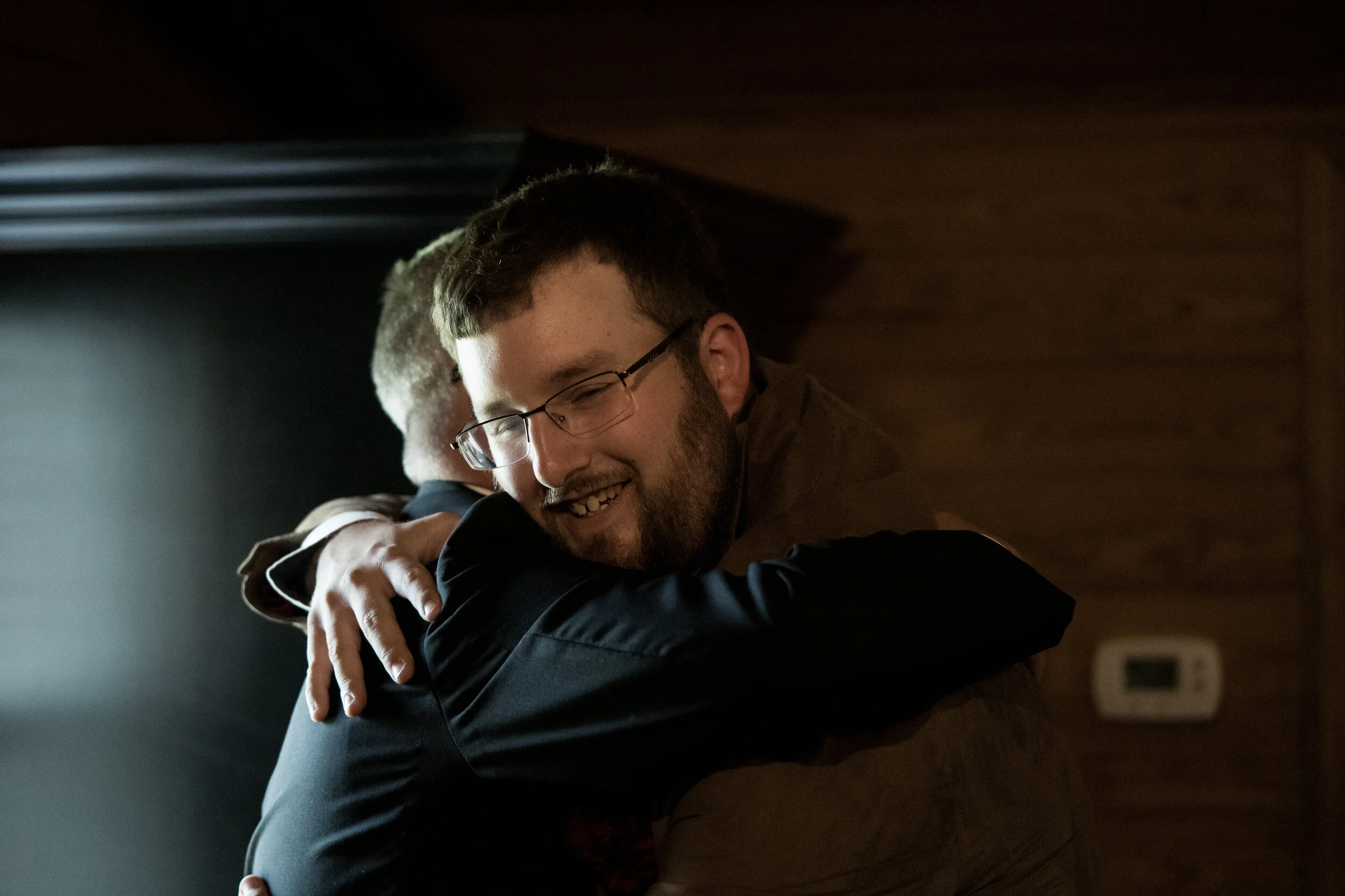 Two men hugging, one smiling with glasses, the other with short hair, in a warmly lit room with wooden walls.