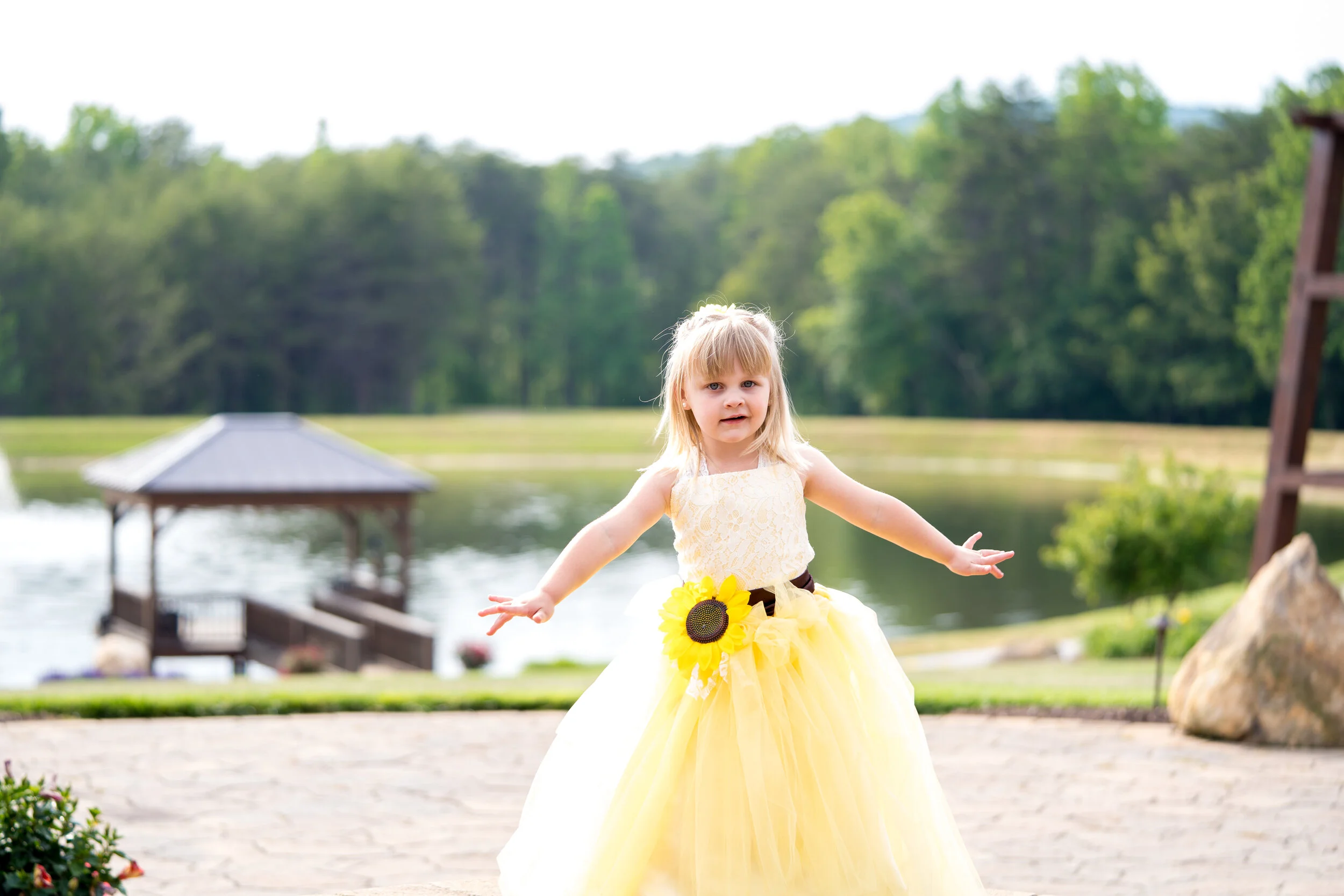 Young girl in a yellow princess dress standing outdoors near a lake, with trees in the background and a wooden gazebo.