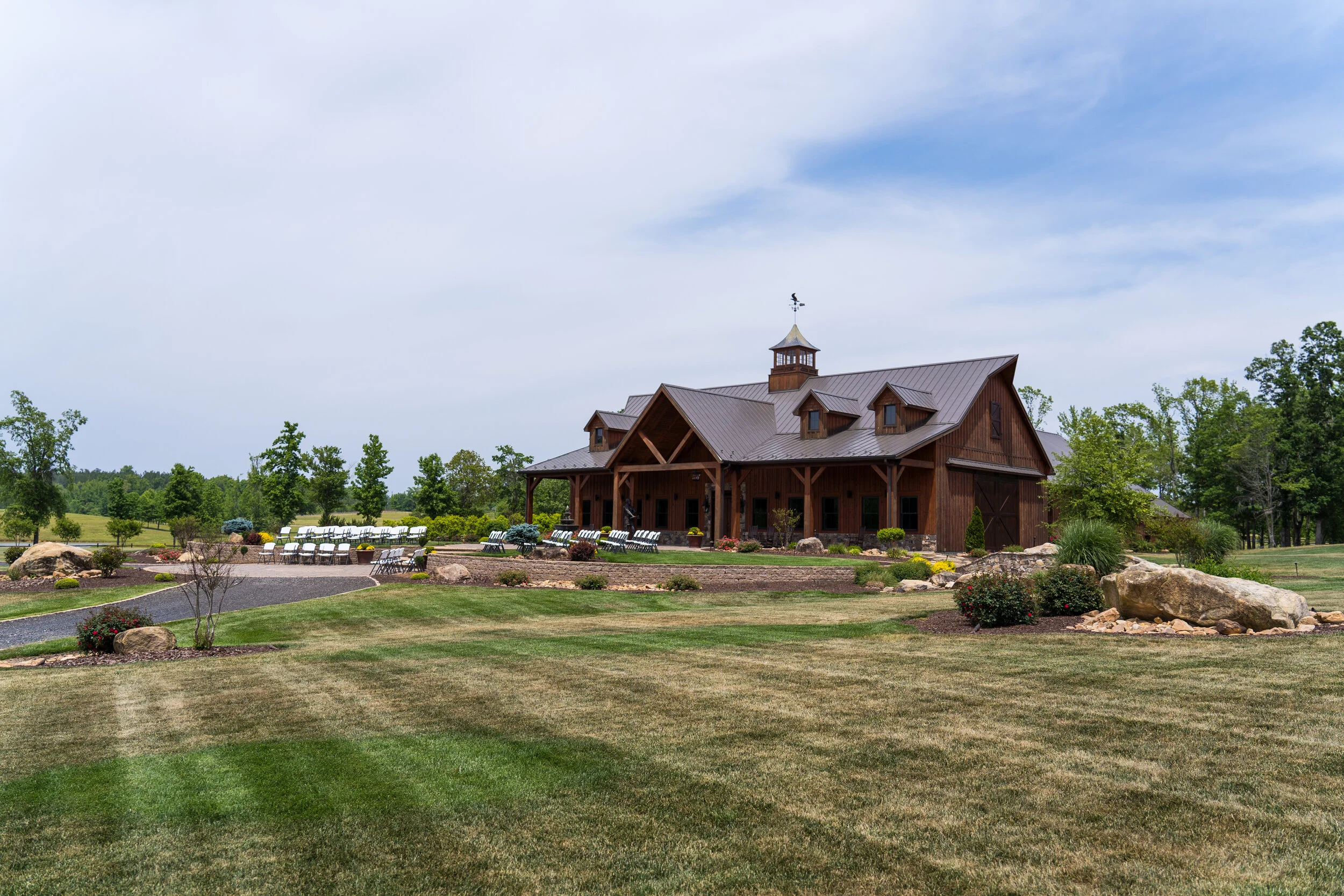 A large wooden barn with a metal roof, surrounded by landscaped green lawns, small trees, rocks, and gardens, under a partly cloudy sky.
