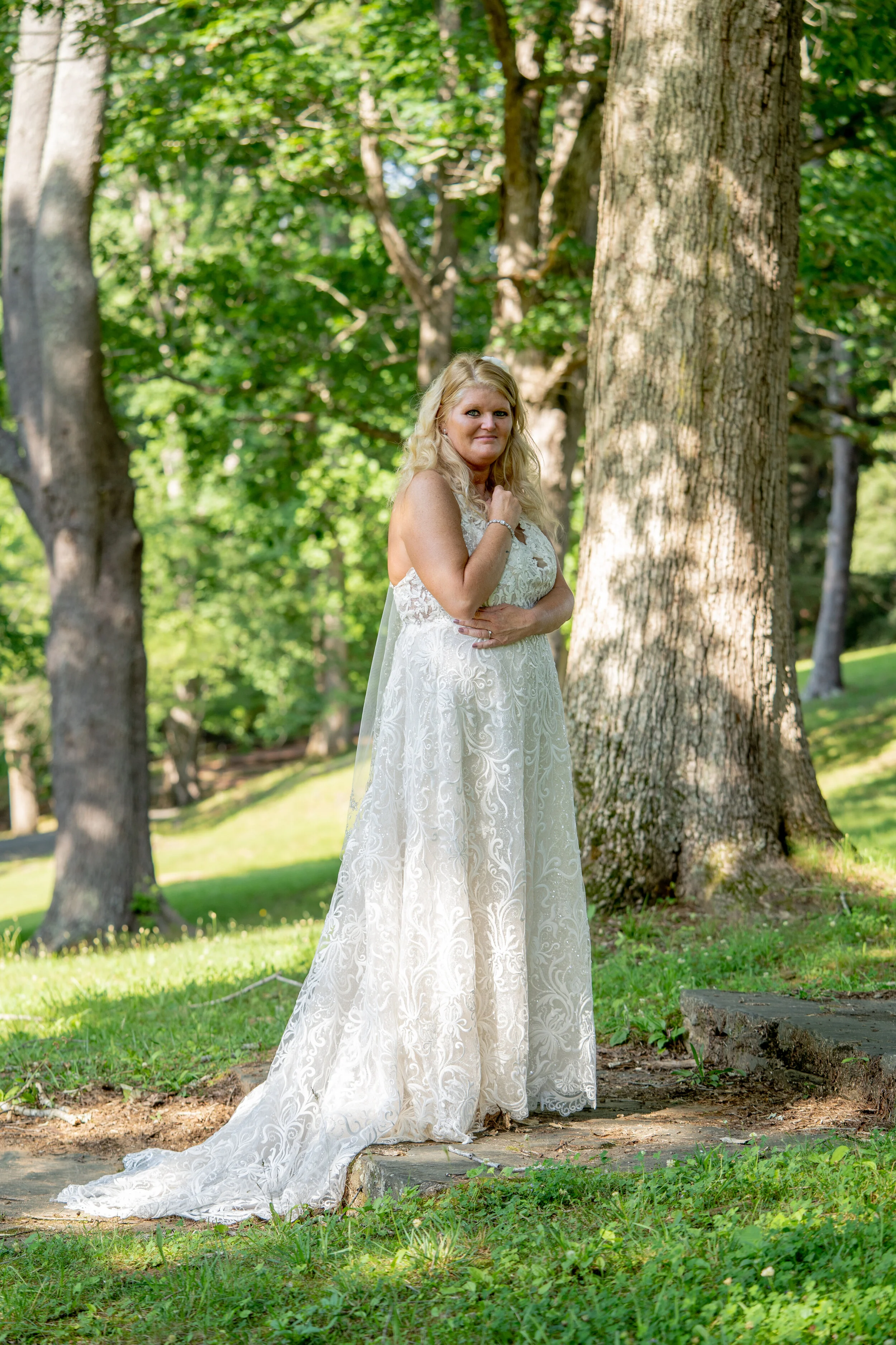 A woman in a white lace wedding gown stands outdoors on a grassy area near trees, with sunlight filtering through the leaves.