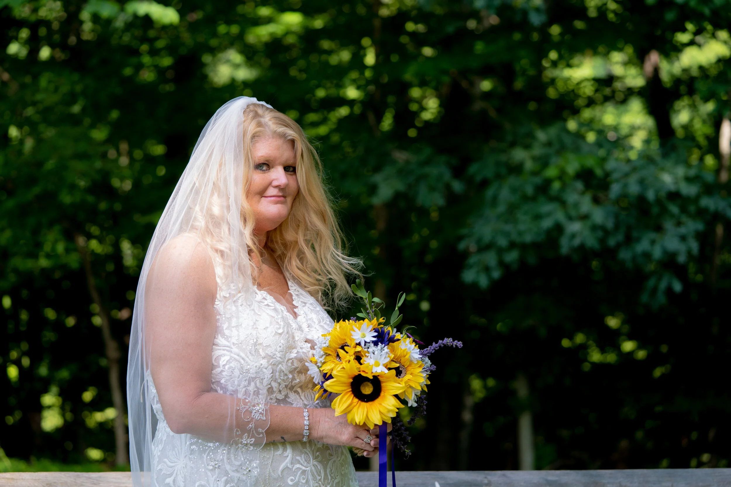 Woman in white wedding dress holding a bouquet of sunflowers, daisies, and purple flowers, standing outdoors in front of green trees, with a white veil on her head.