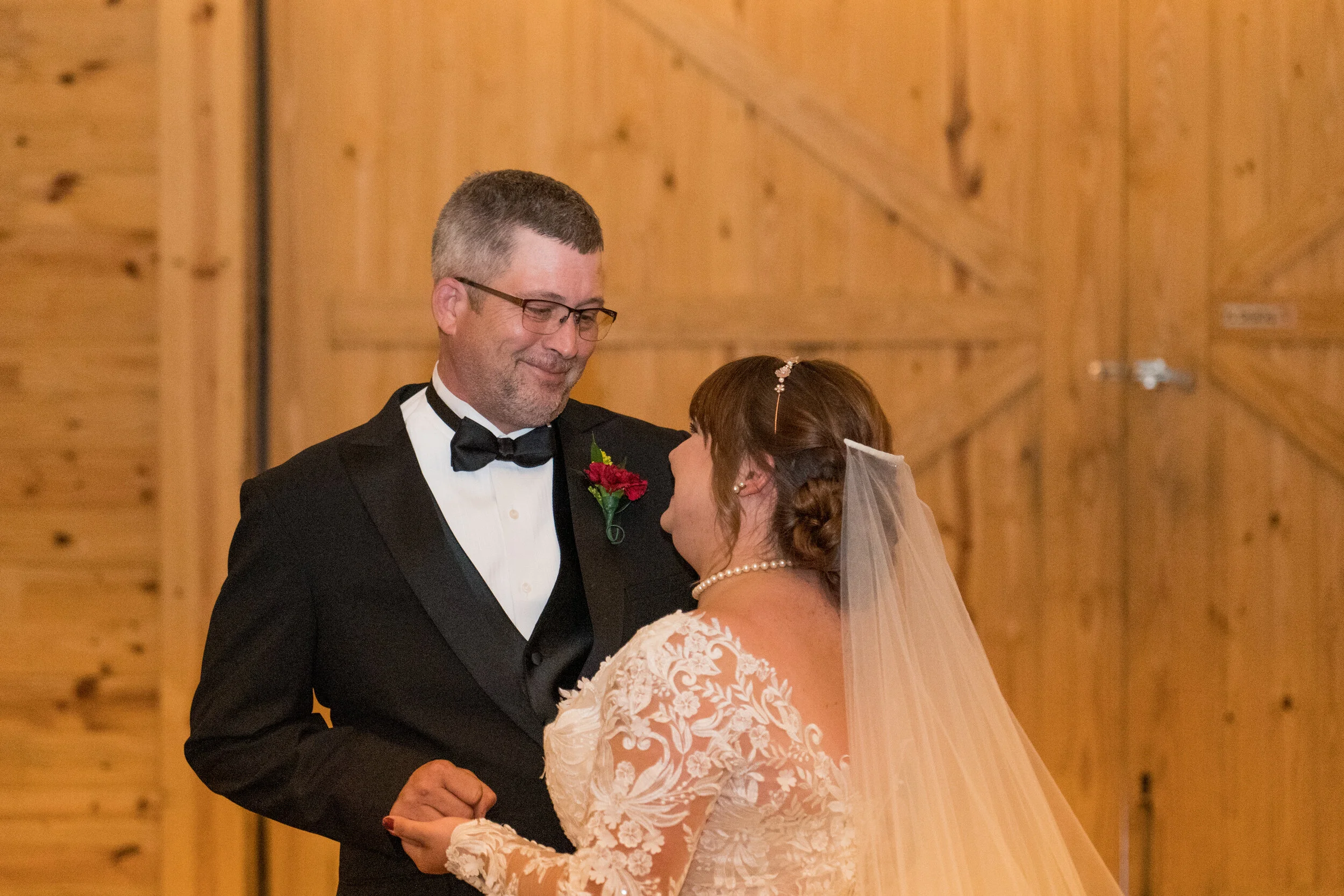A groom in a black tuxedo and a bride in a lace wedding dress and veil dance together in a wooden hall.