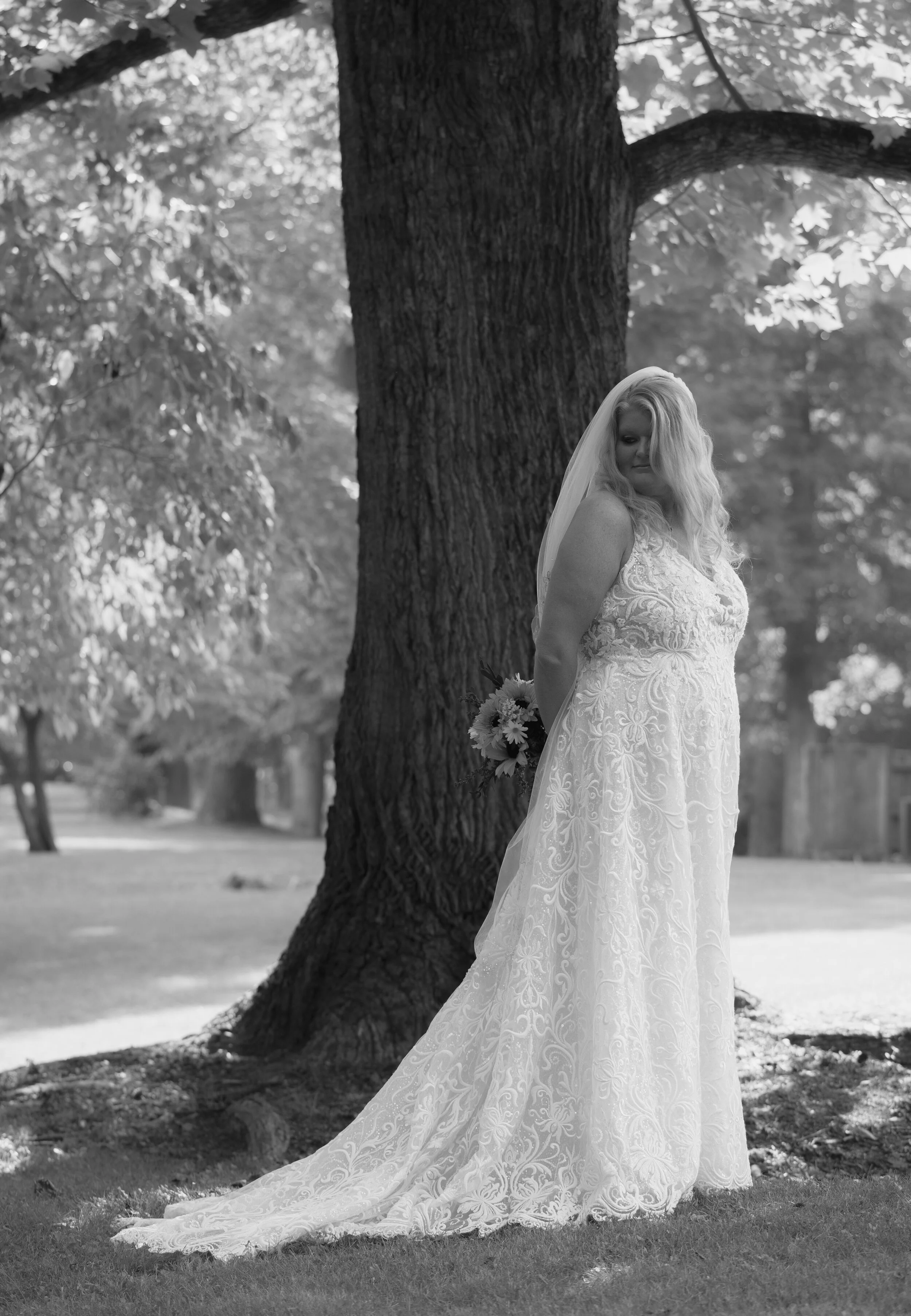 A bride in a lace wedding gown and veil stands outdoors by a large tree, holding a bouquet of flowers behind her back, in black and white.
