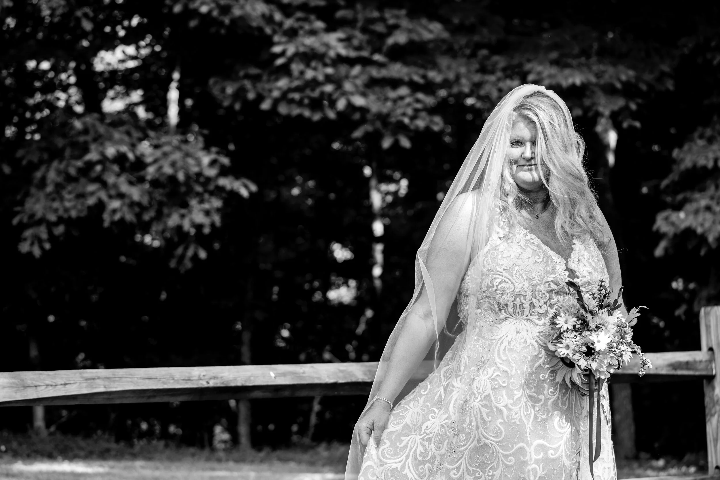 Black and white photograph of a bride standing outdoors, wearing a detailed lace wedding dress and a veil, holding a bouquet of flowers, with trees and a wooden fence in the background.