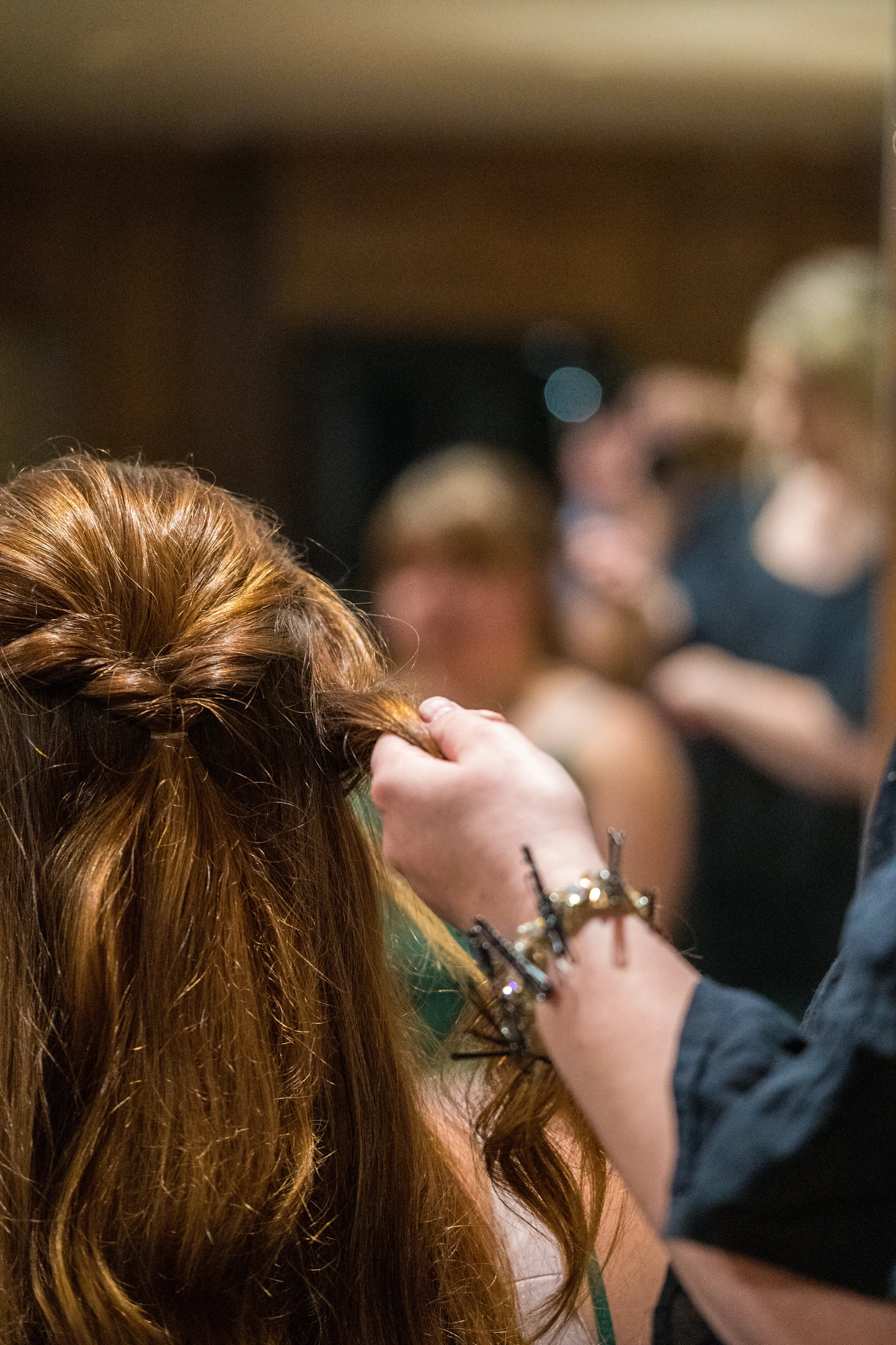 A hairstylist working on a woman's red hair at a salon or event, with blurred people in the background.