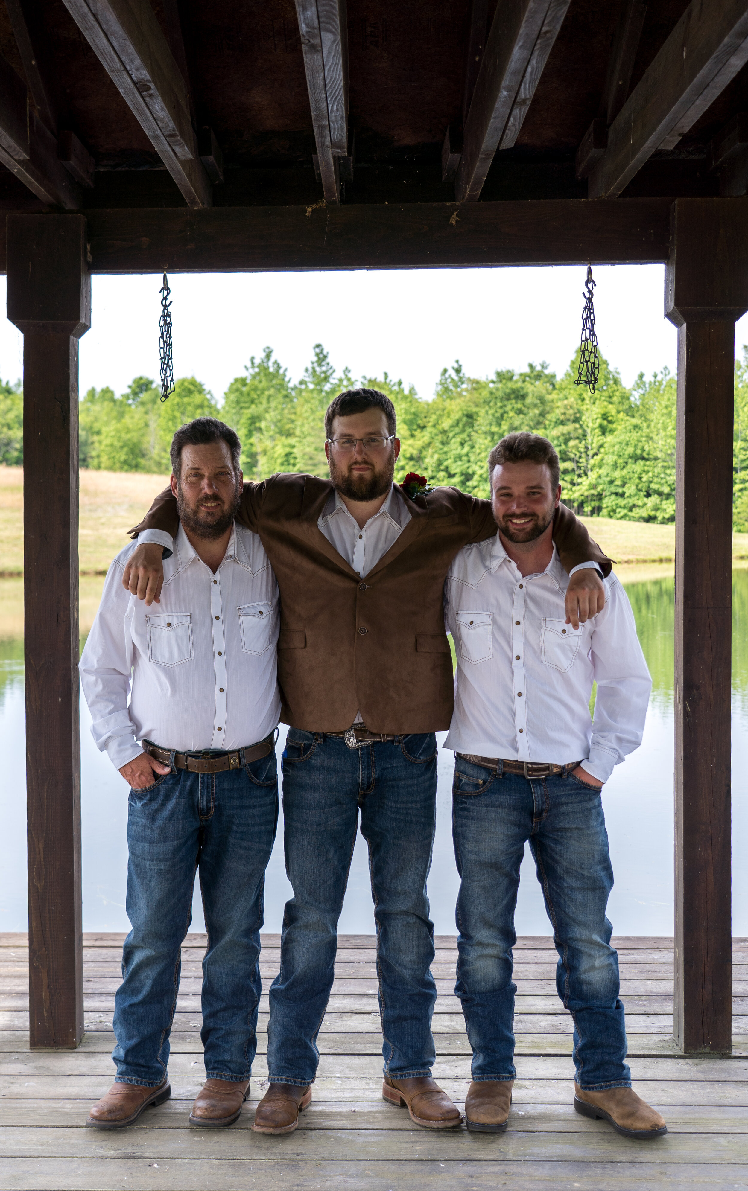 Three men standing side by side outdoors under a wooden structure, with a lake and green trees in the background. The man in the middle wears a brown jacket, while the two on either side wear white shirts and jeans. All have their arms around each ot