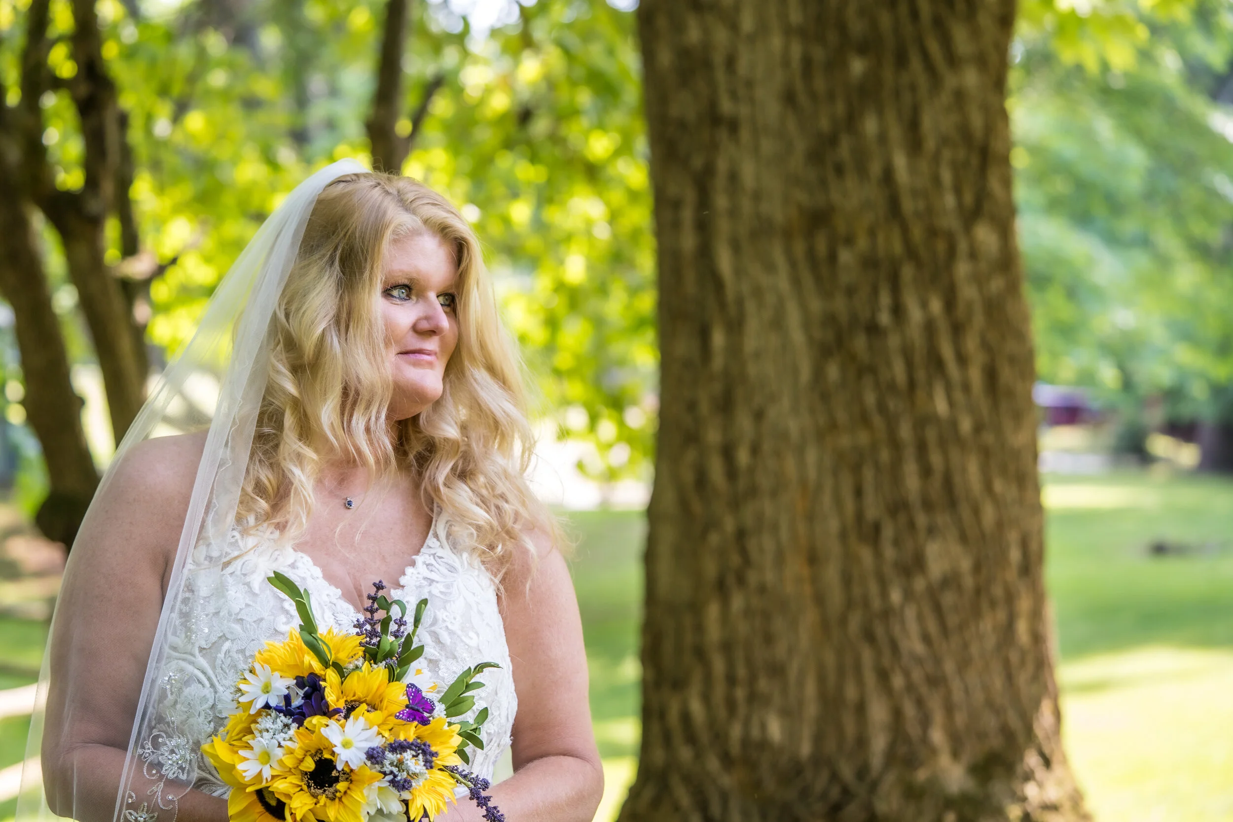 A bride with long blonde hair wearing a white lace wedding gown and veil, holding a bouquet of yellow and purple flowers, standing outdoors near a large tree in a park.