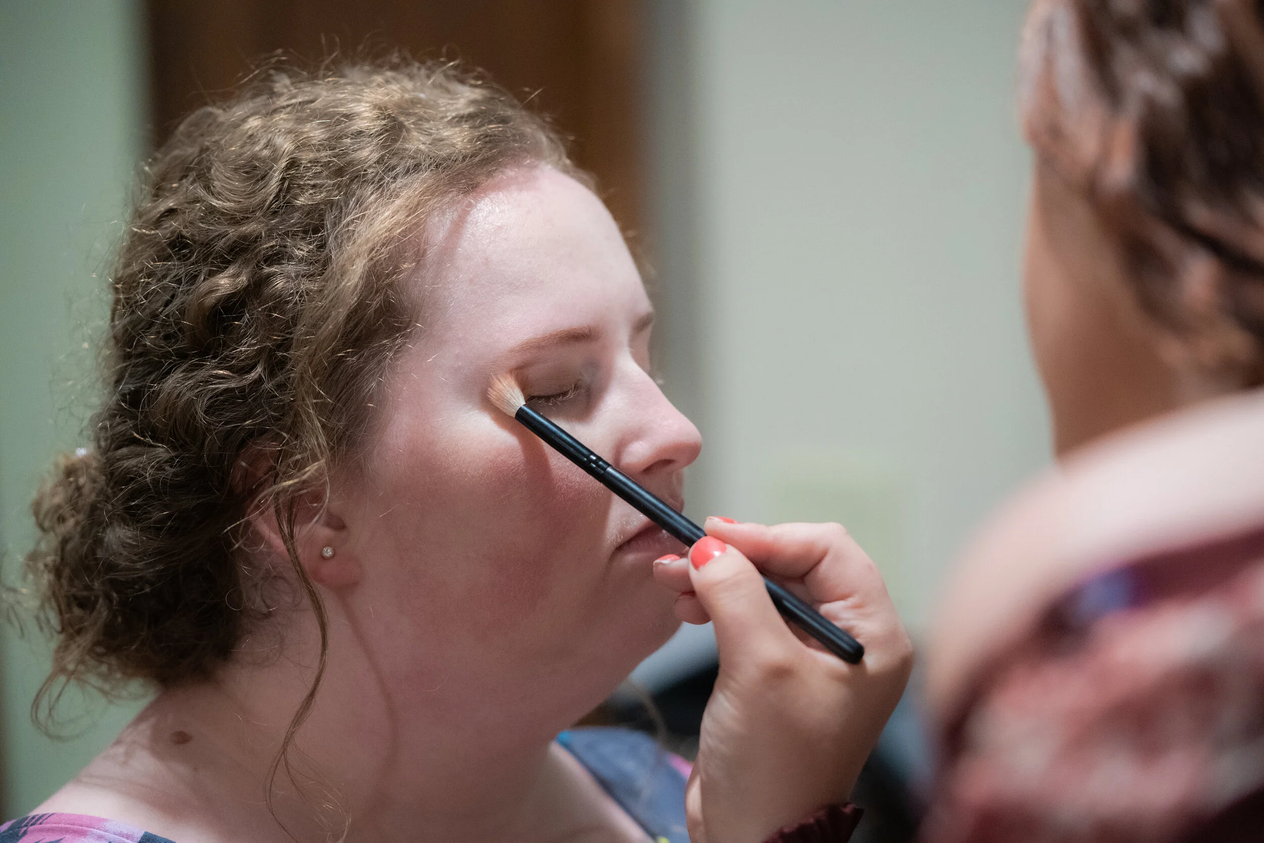 A woman with curly blonde hair getting her eyeshadow applied by a makeup artist.