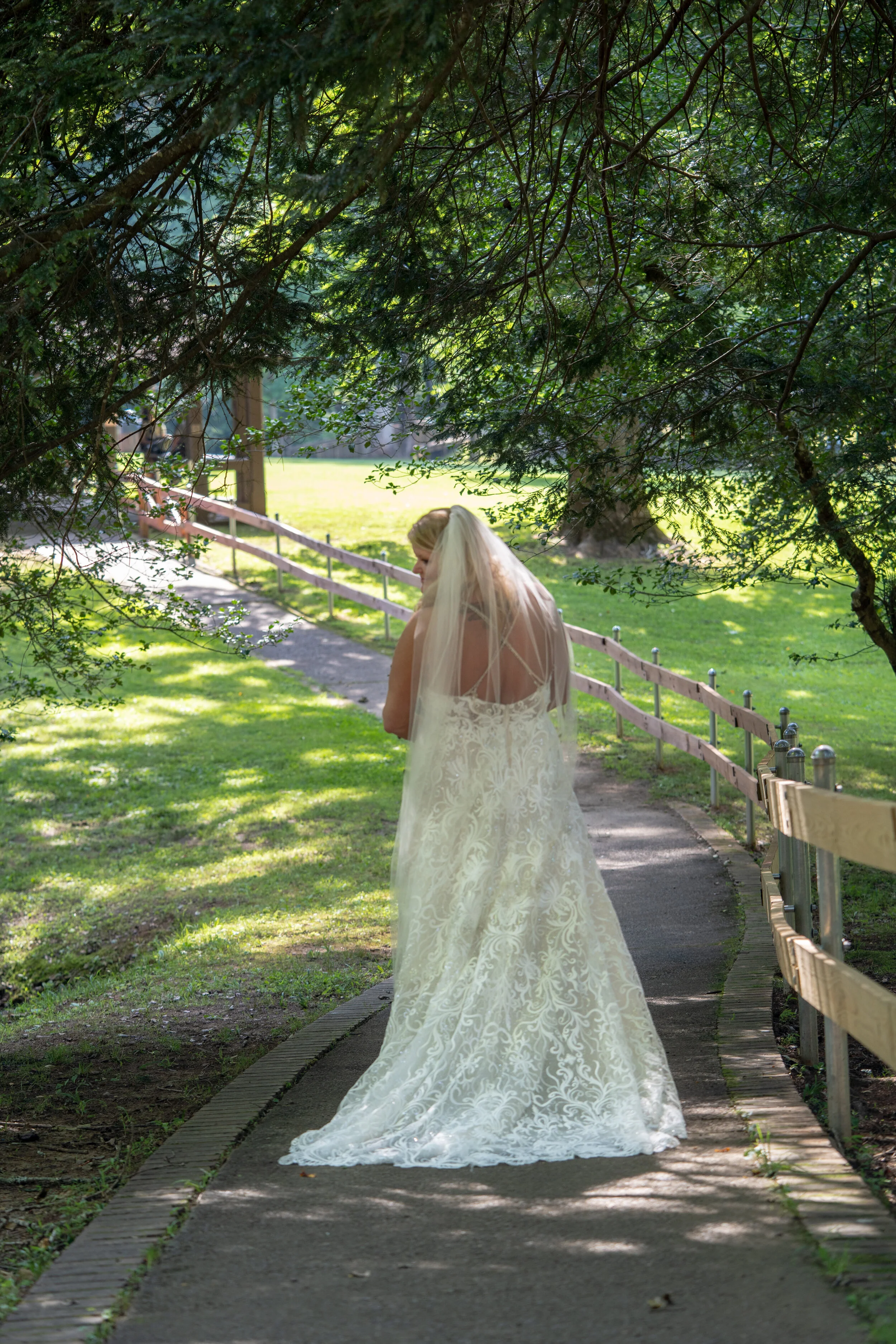 Bride in a white wedding gown and veil walking on a paved path through a green park, shaded by trees.