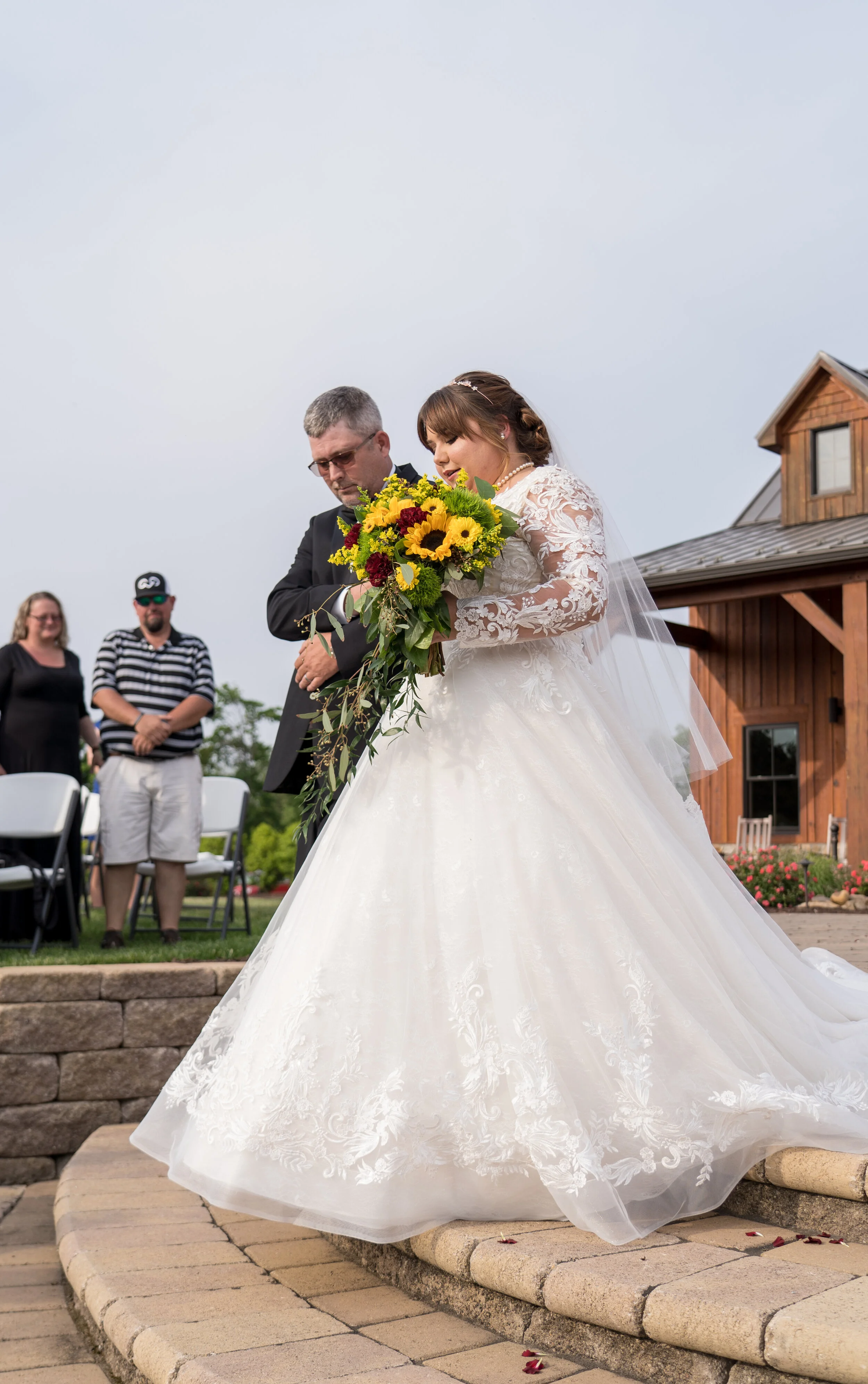 A bride in a white wedding gown holding a bouquet of sunflowers and red flowers, walking with a man outside during a wedding ceremony, with guests and a wooden building in the background.