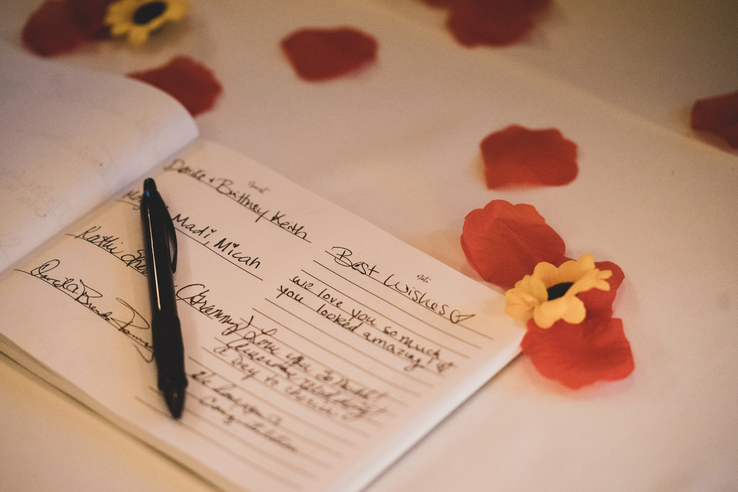 Open guest book with handwritten notes and a black pen resting on it, decorated with red and yellow flower petals scattered around on a white surface.
