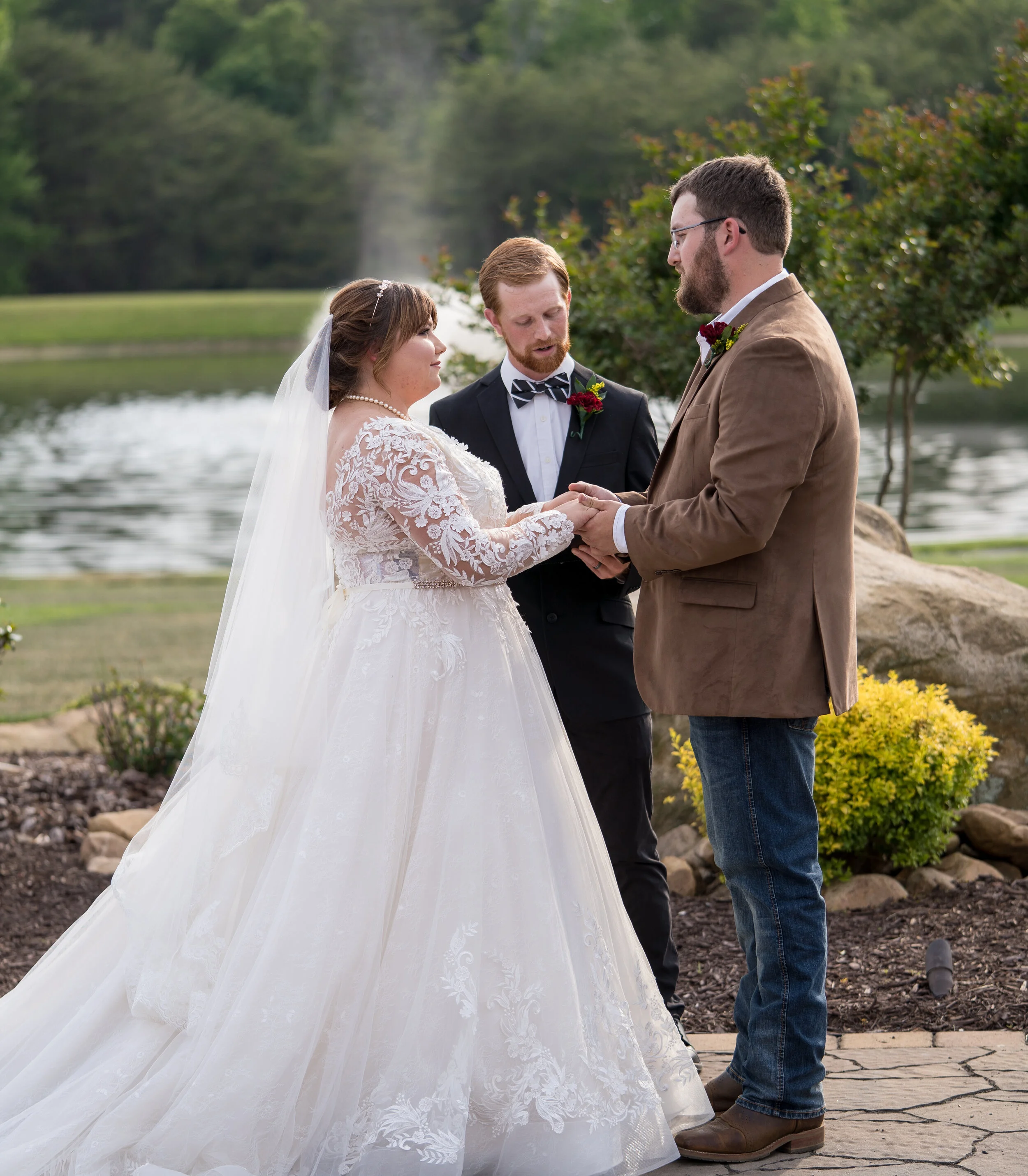 Bride and groom exchanging wedding vows outdoors near a lake with officiant officiating, trees, rocks, and yellow bushes in the background.