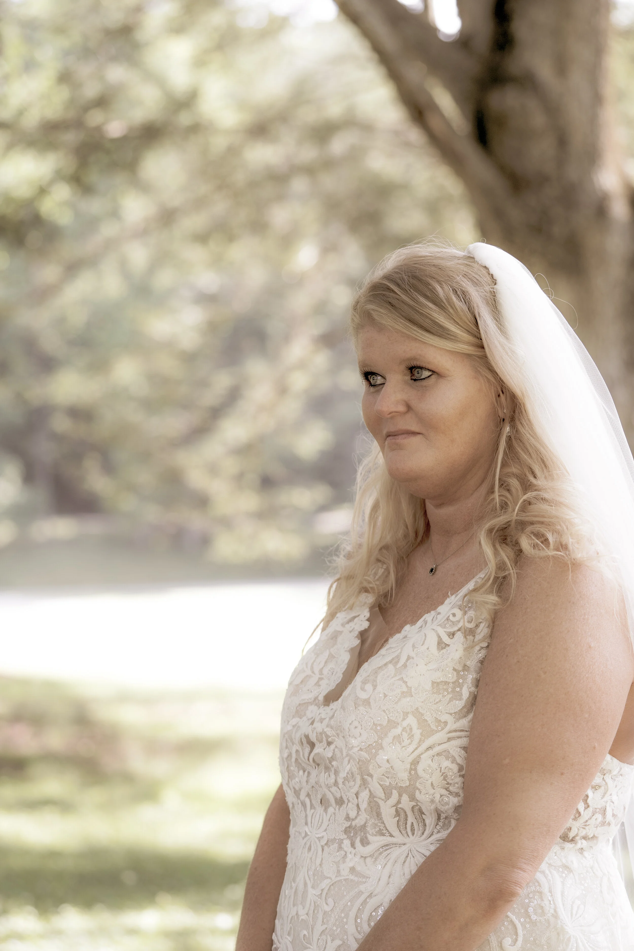 Bride in a lace wedding dress with a veil, standing outdoors near a tree.