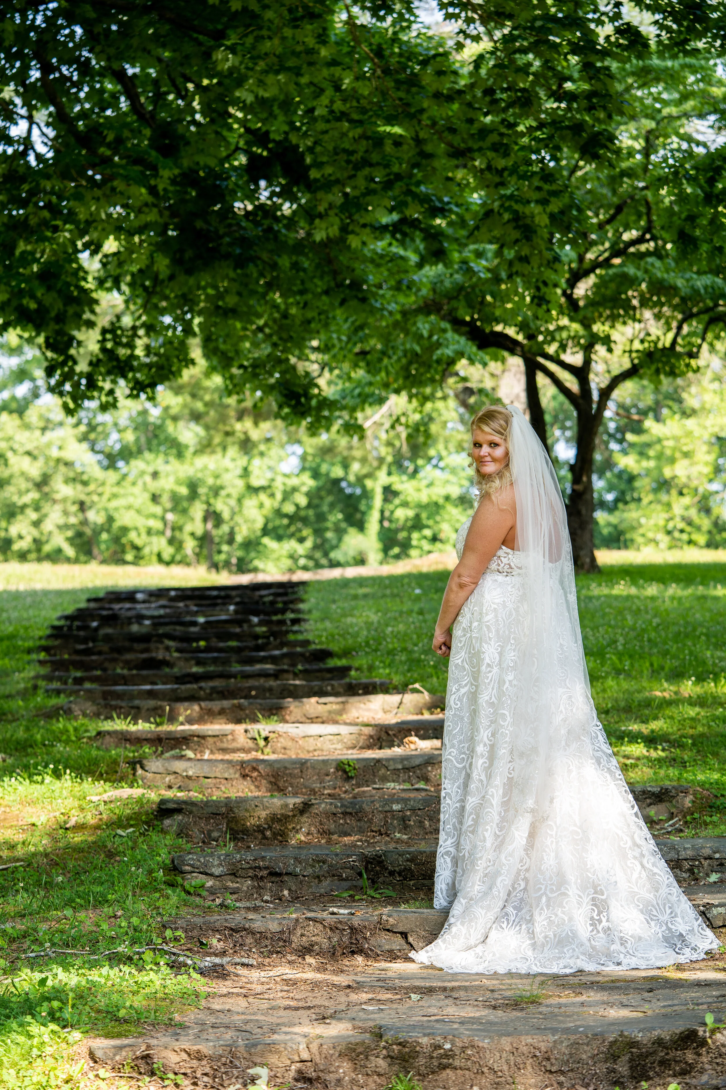 Bride in a white wedding dress standing on outdoor stone steps under a large leafy tree.