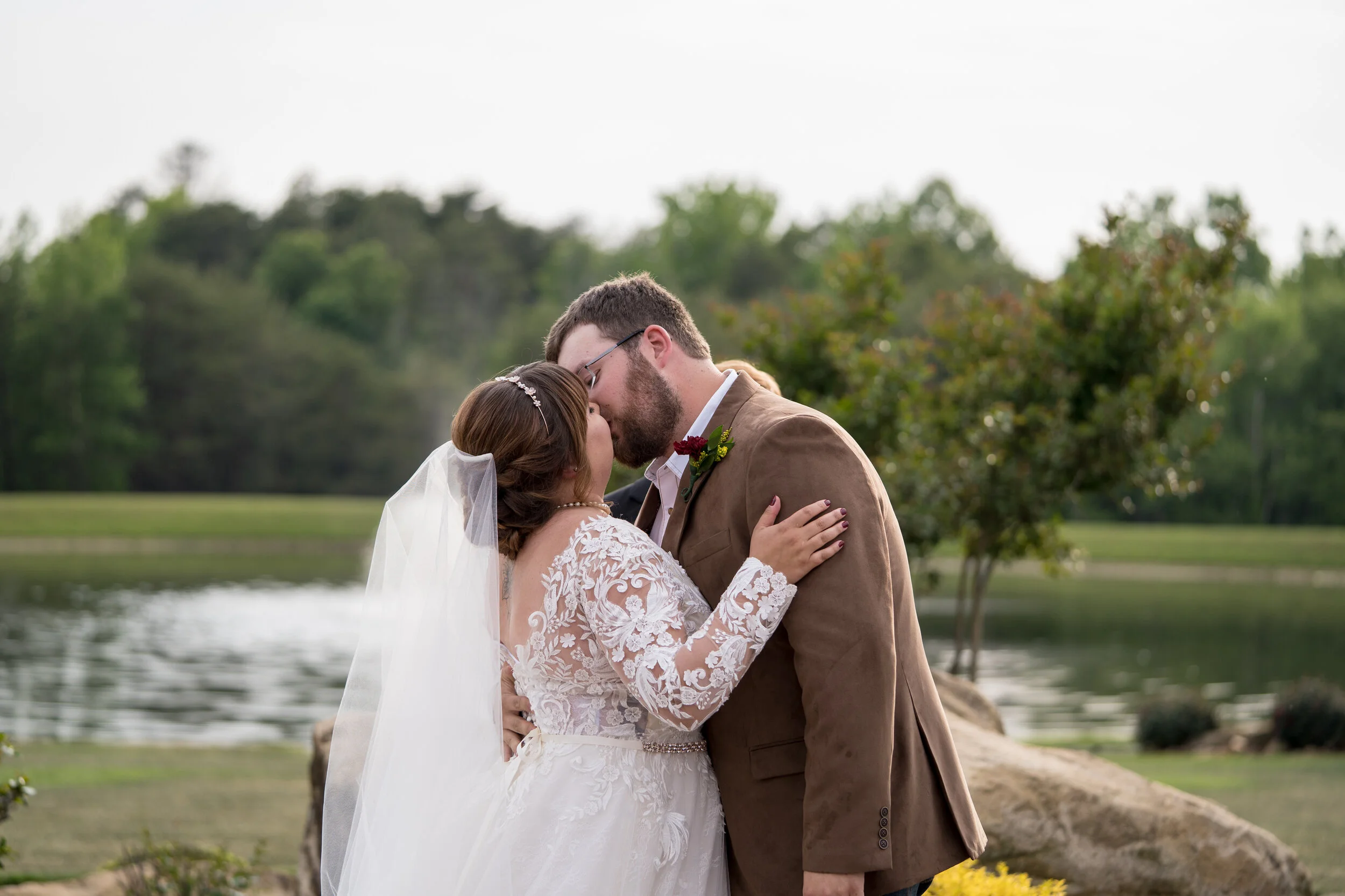 Bride and groom kissing outdoors near a lake with trees in the background.