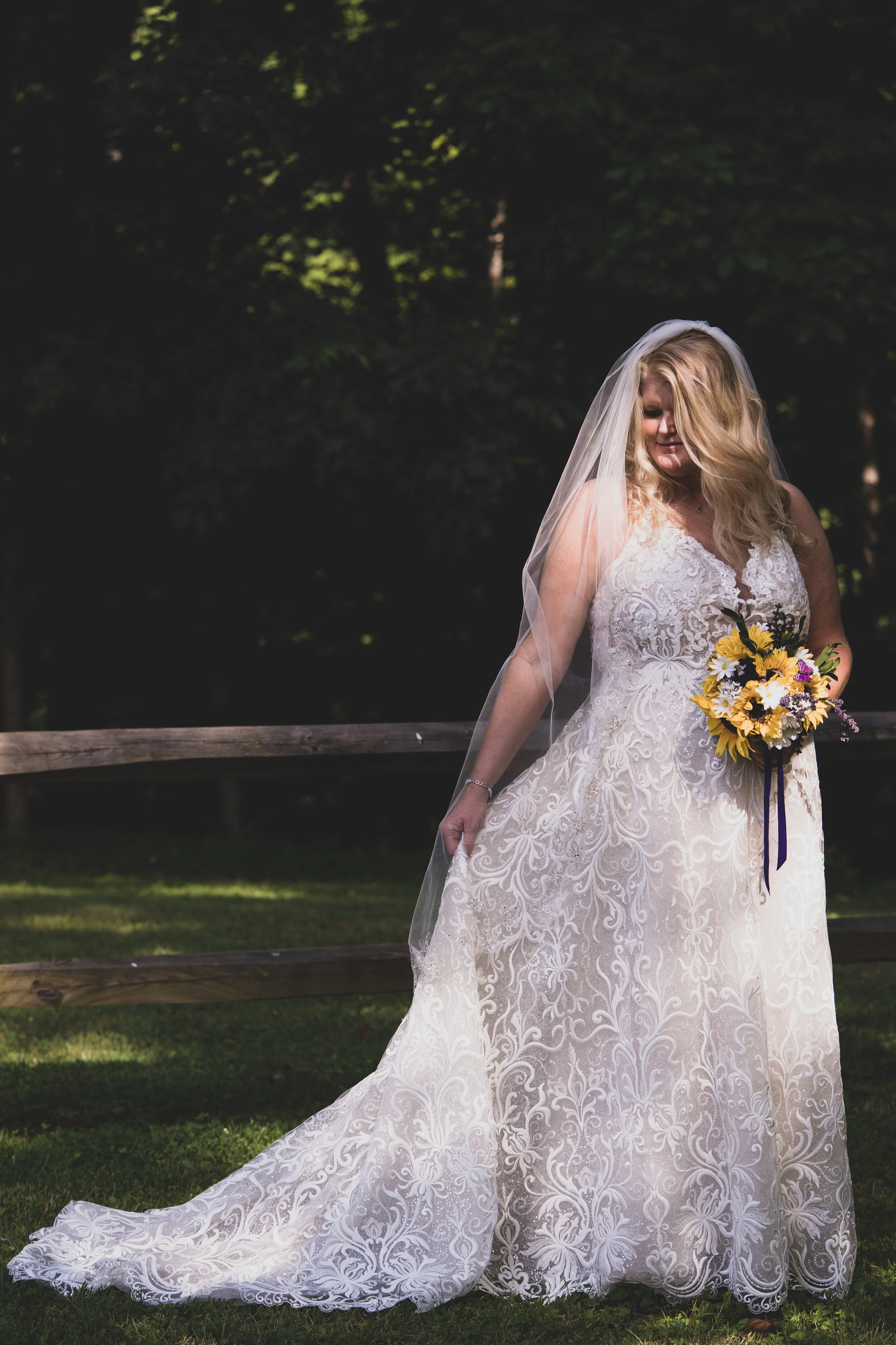 A bride in a white lace wedding gown and veil holding a bouquet of yellow and purple flowers outdoors in a wooded area.