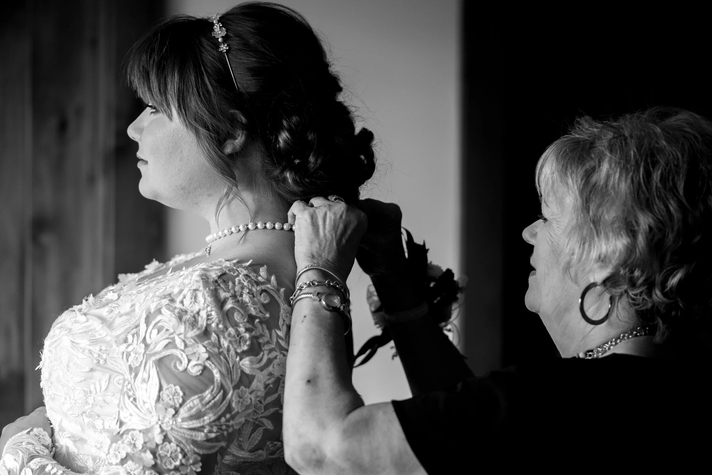 A woman, likely a bride, is being helped with jewelry by an older woman, possibly her mother or a helper. The bride wears an embroidered dress, pearl necklace, and has styled hair with a decorative pin. The older woman is dressed in black, wearing ho