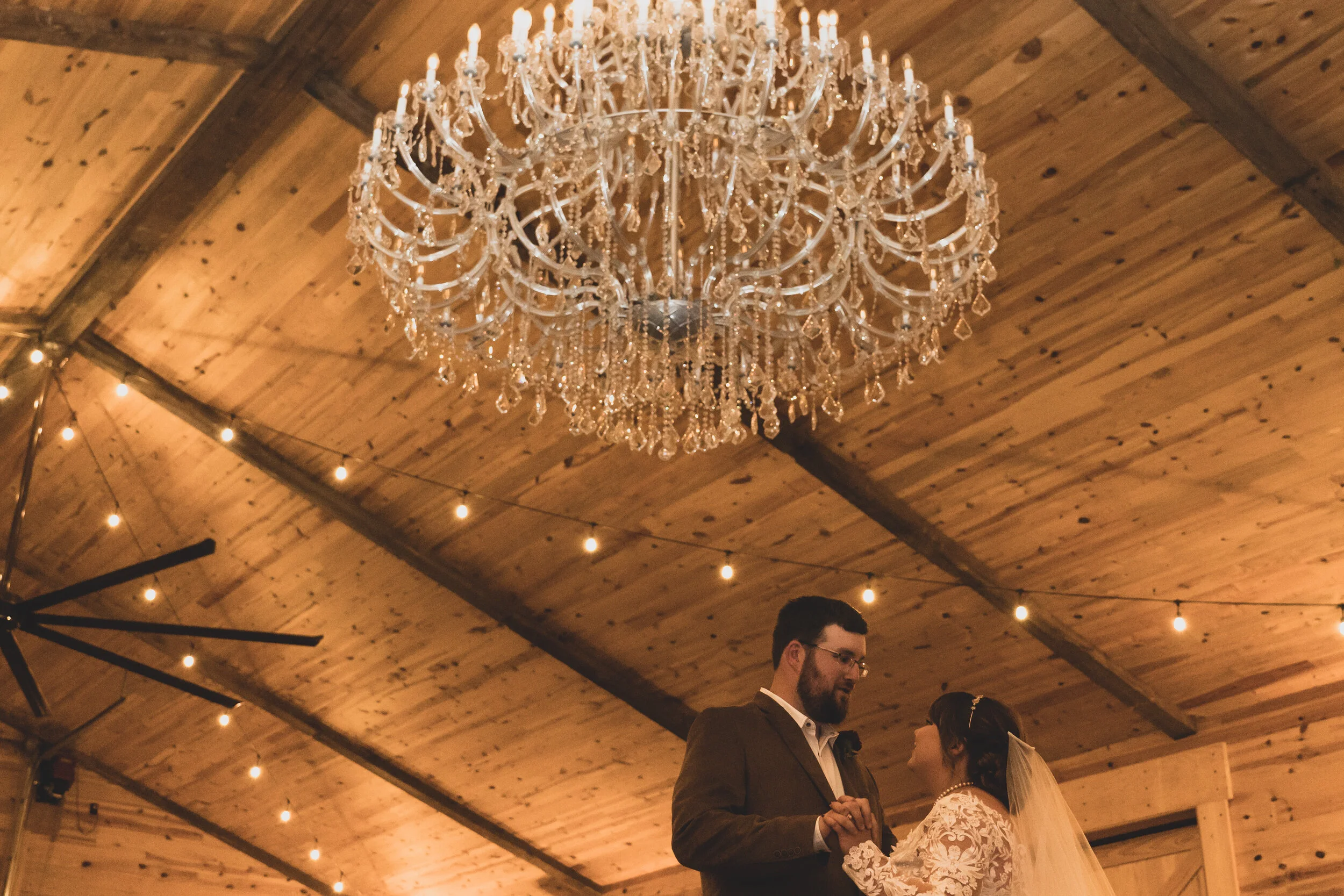 A bride and groom dancing at their wedding reception in a rustic wooden venue with string lights and a large crystal chandelier overhead.