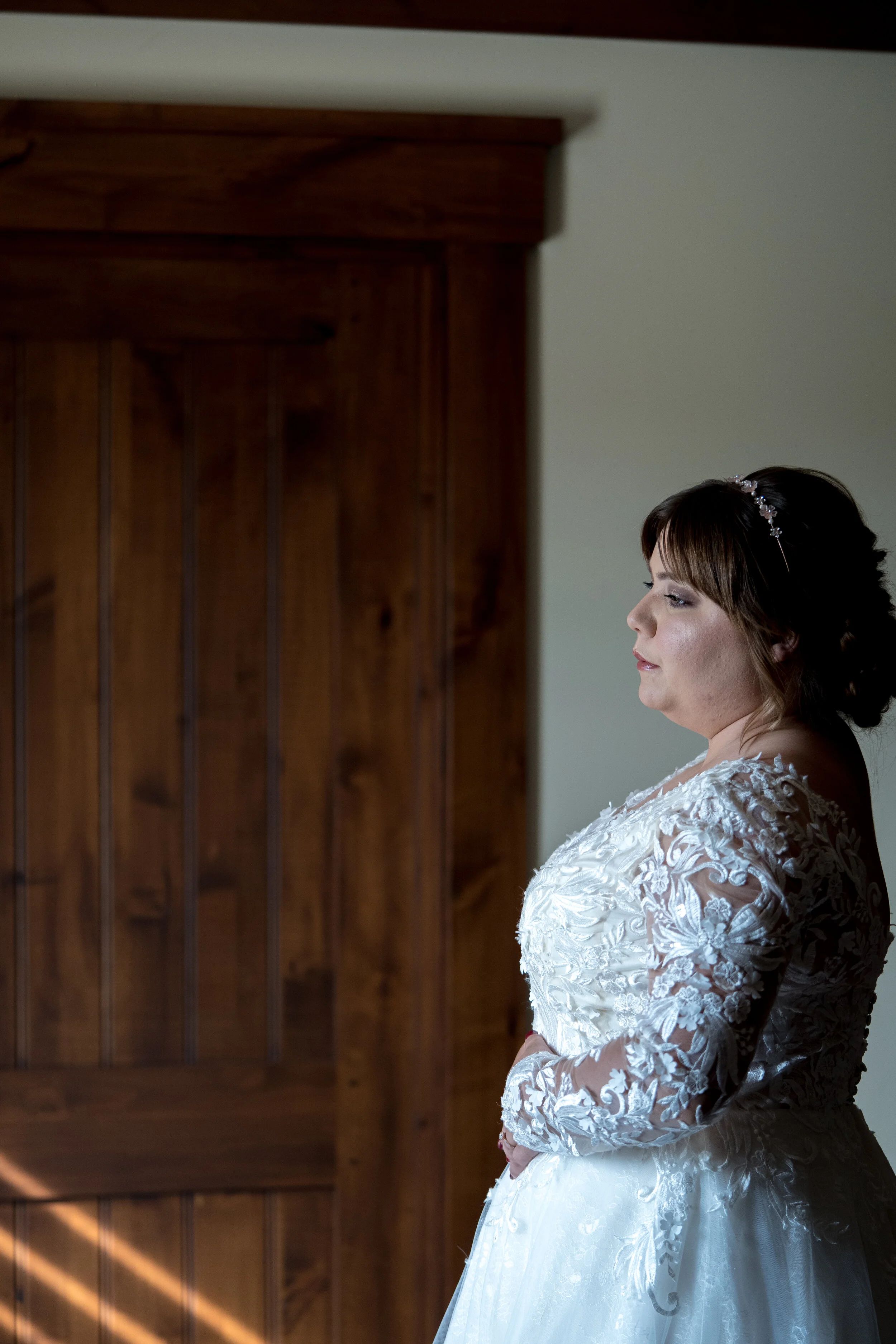 A bride standing indoors, wearing a white lace wedding dress, with her hands folded in front of her, looking to the right.