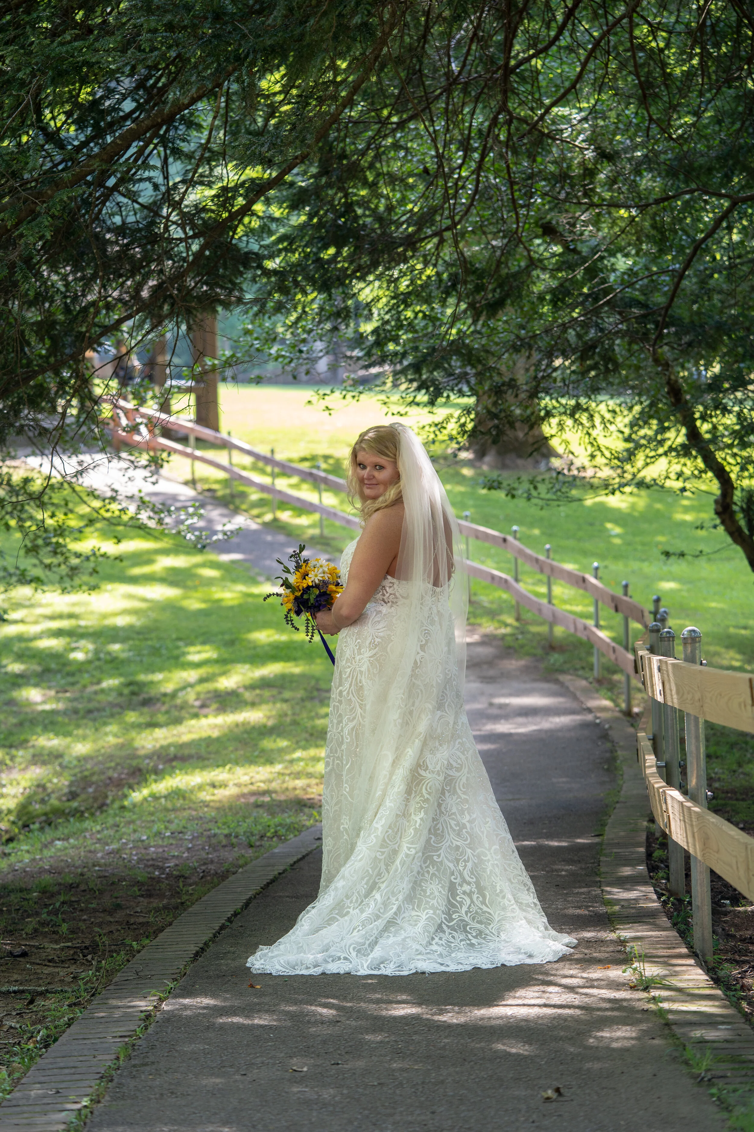 A bride in a white wedding dress holding a bouquet of yellow and purple flowers walking along a shaded garden path.