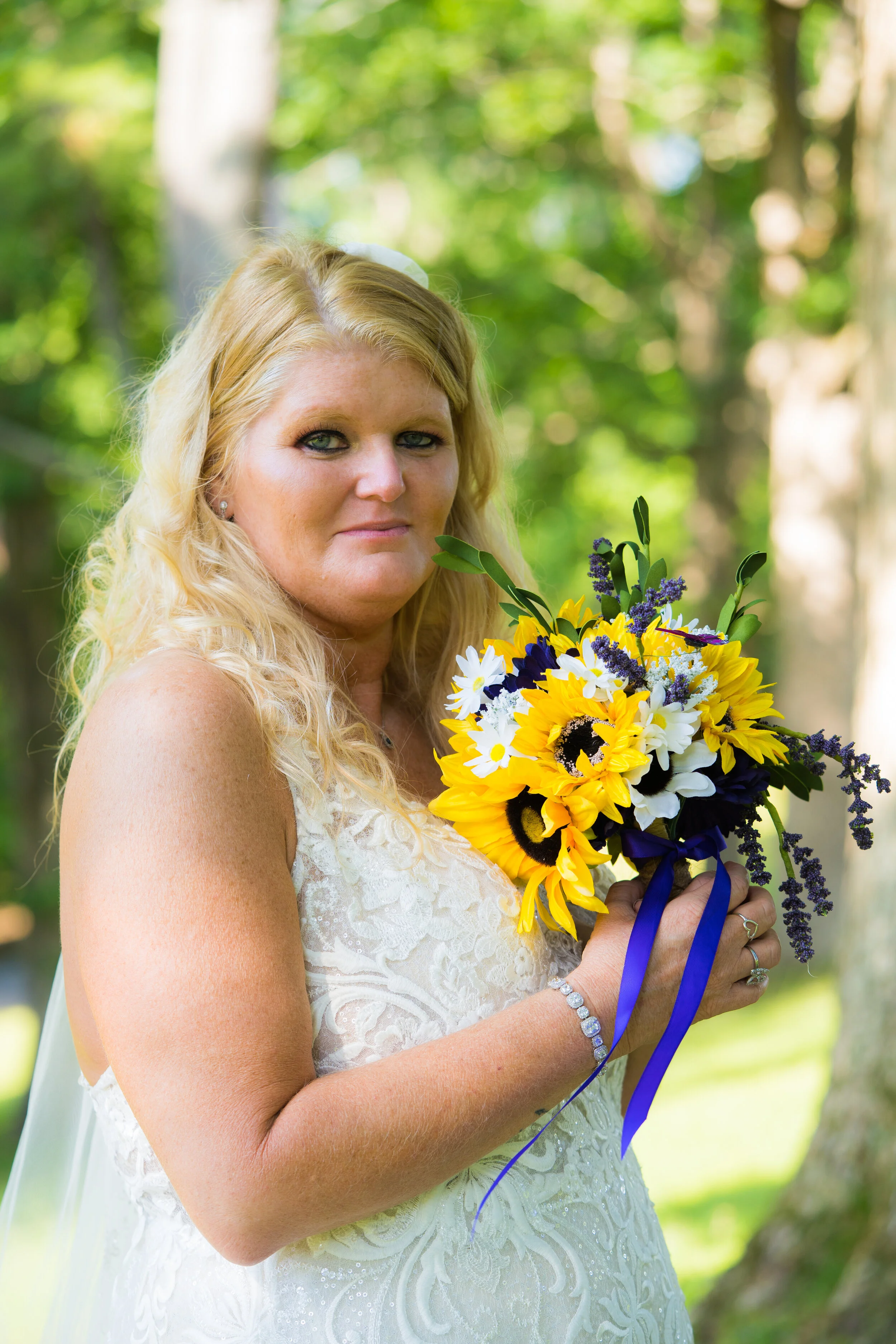 A woman in a white lace dress holding a bouquet of sunflowers, daisies, and purple flowers outdoors with a green, leafy background.