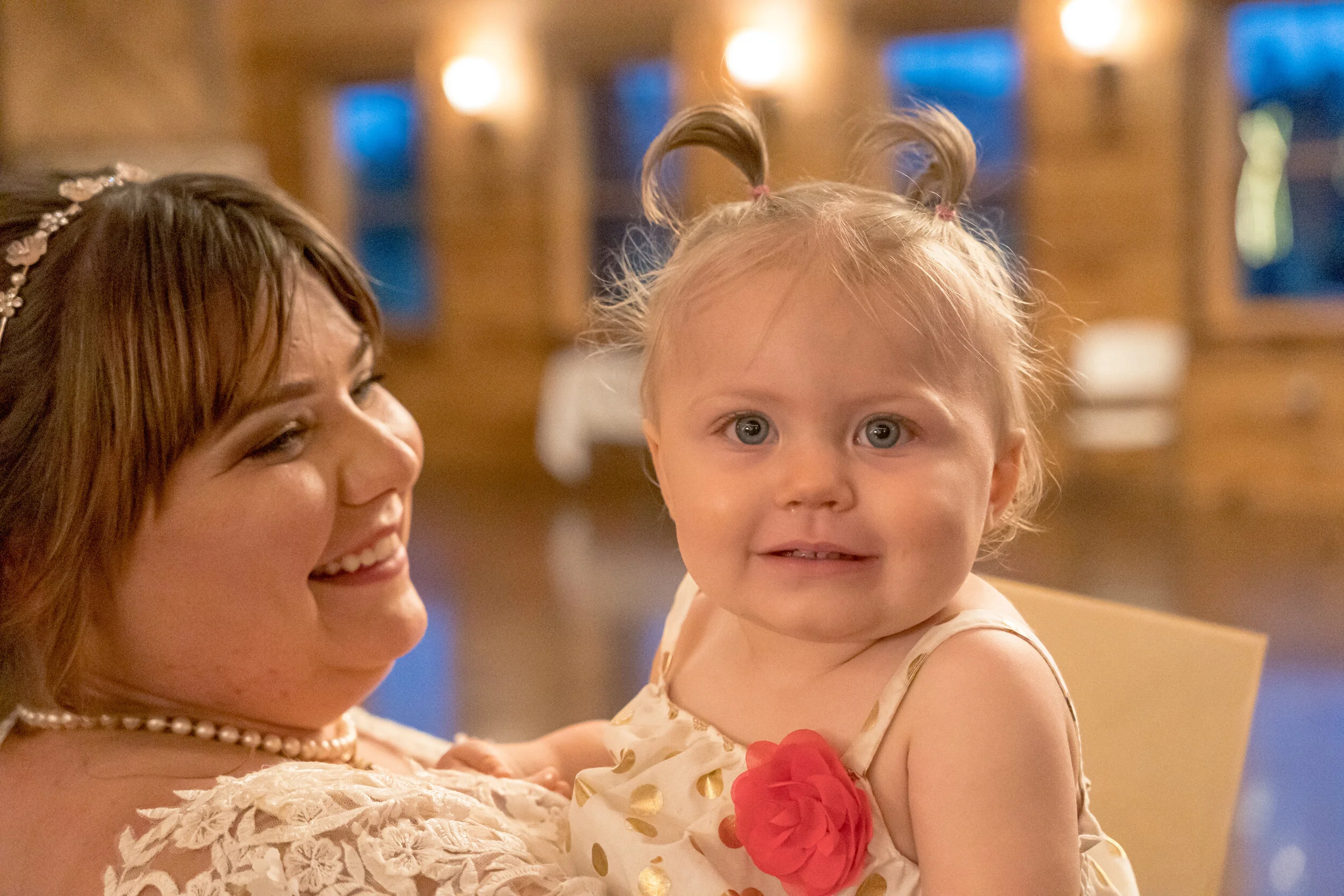 A smiling woman holding a young girl with pigtails in a warmly lit indoor setting.