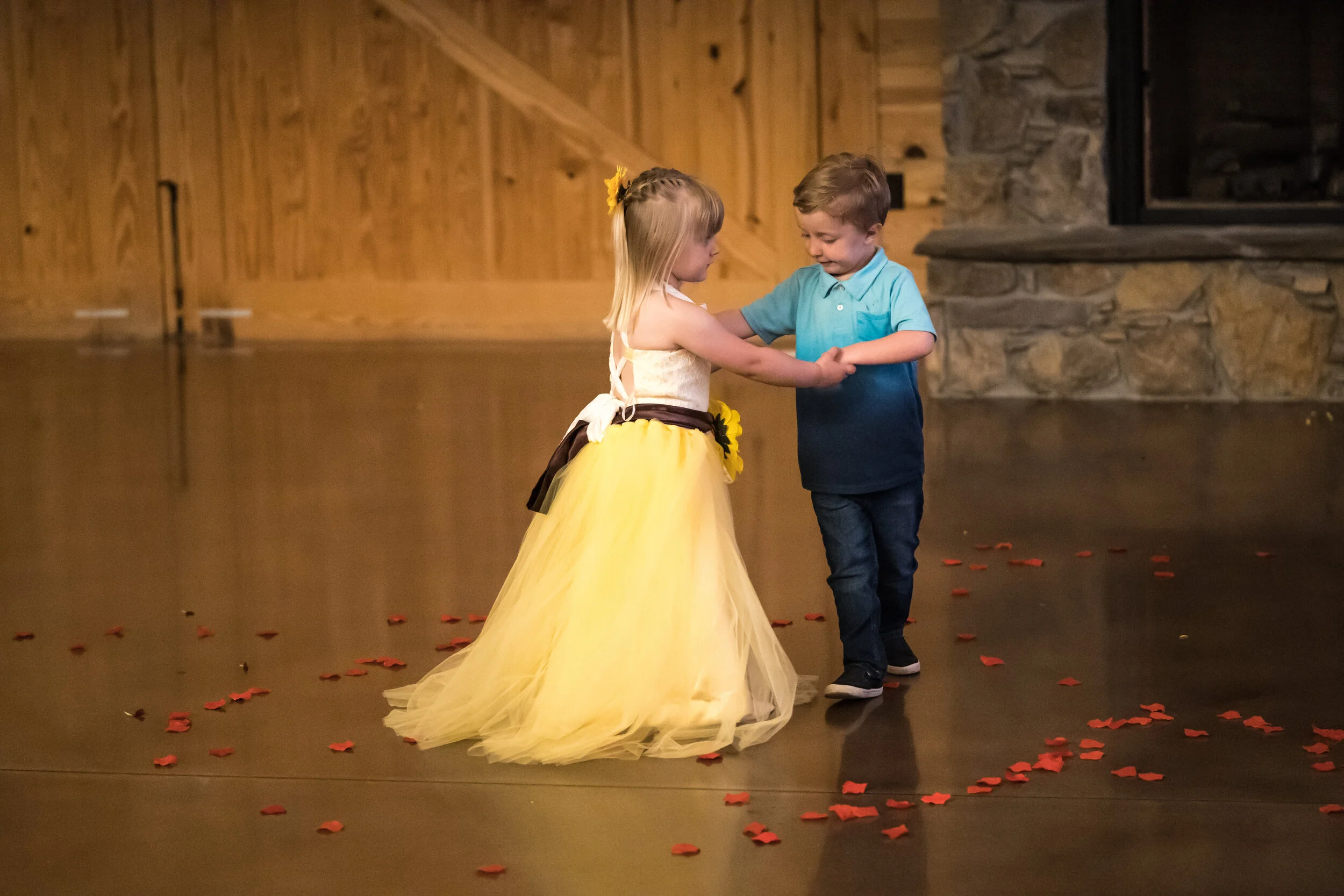 Two young children dancing together in a wooden-finished room. The girl is wearing a yellow dress with a flower detail, and the boy is dressed in a blue shirt and dark jeans. They are holding hands and smiling.