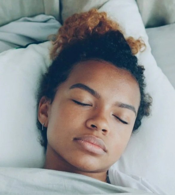 Young woman with curly hair lying in bed with eyes closed, appearing to be resting or sleeping.