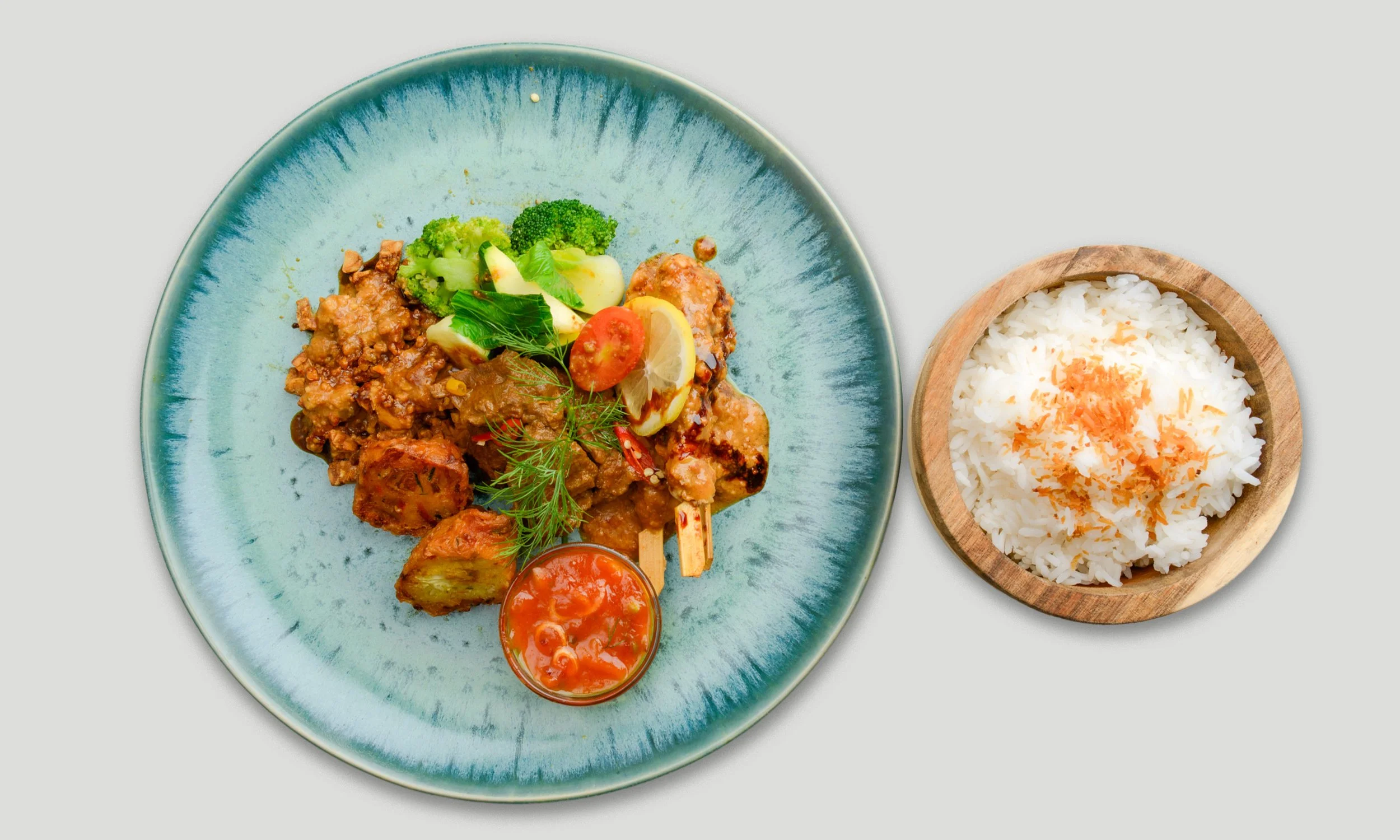 Ein Teller mit indischem Curry mit Gemüse und Fleisch, Garnele, Kirschtomaten, Zitronenstücken und frischen Kräutern, daneben eine Schüssel mit weißem Reis, garniert mit geröstetem Sesam.