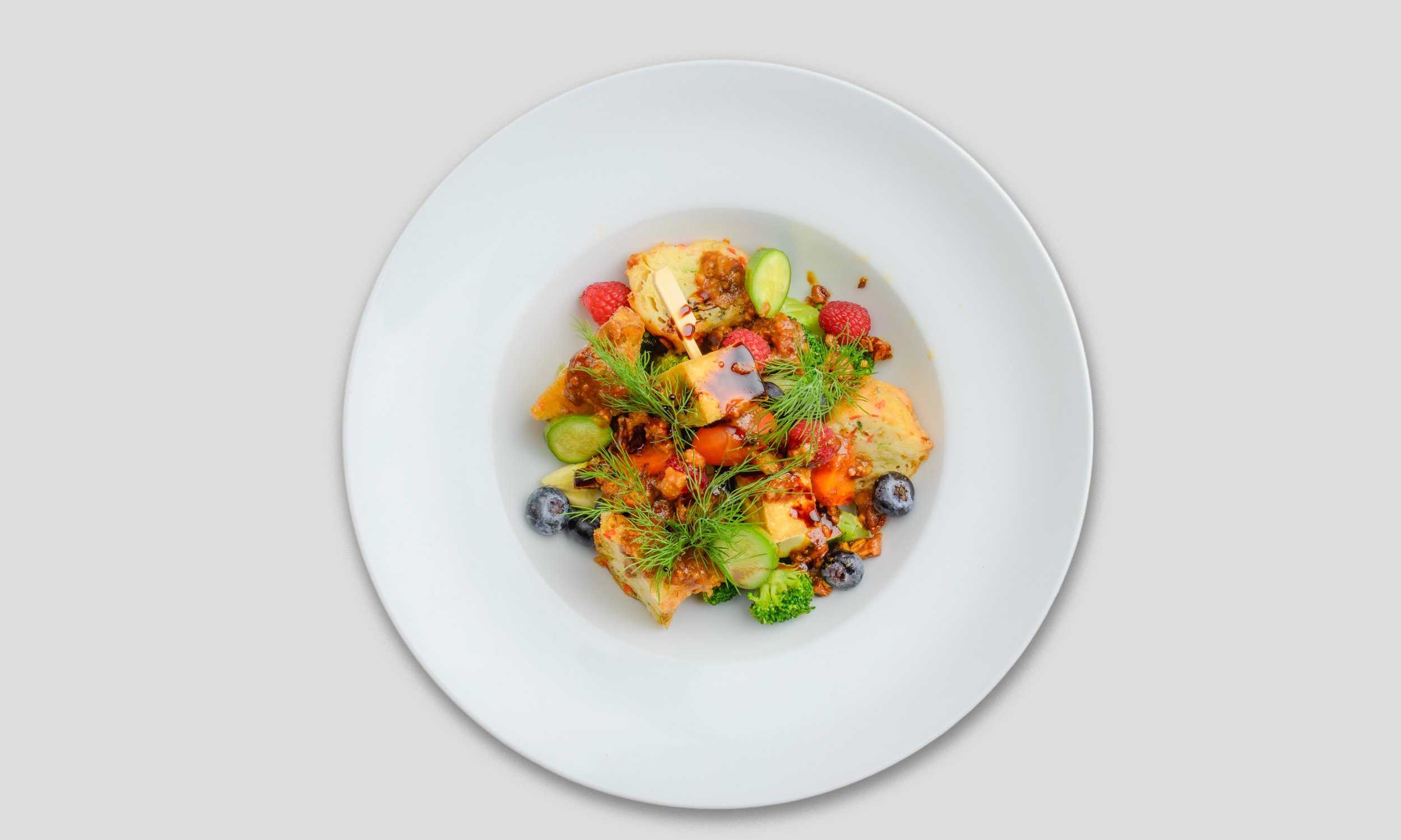 Ein Teller mit einem bunten Gericht aus Kartoffelauflauf, frischen Beeren, Gurken, Minz- und Dill-Garnitur, und einer dunklen Soße