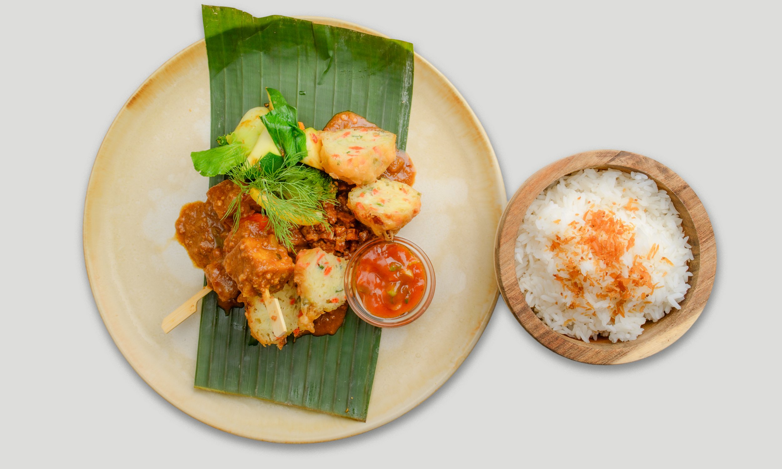 Ein Teller mit verschiedenen indischen Gerichten, darunter vegetarische Köttbullar, Gemüseteller, eine kleine Schüssel mit rotem Chutney und ein kleiner Holzteller mit weißem Reis, serviert auf einem ovalen Keramikteller mit einer grünen Bananenblatt