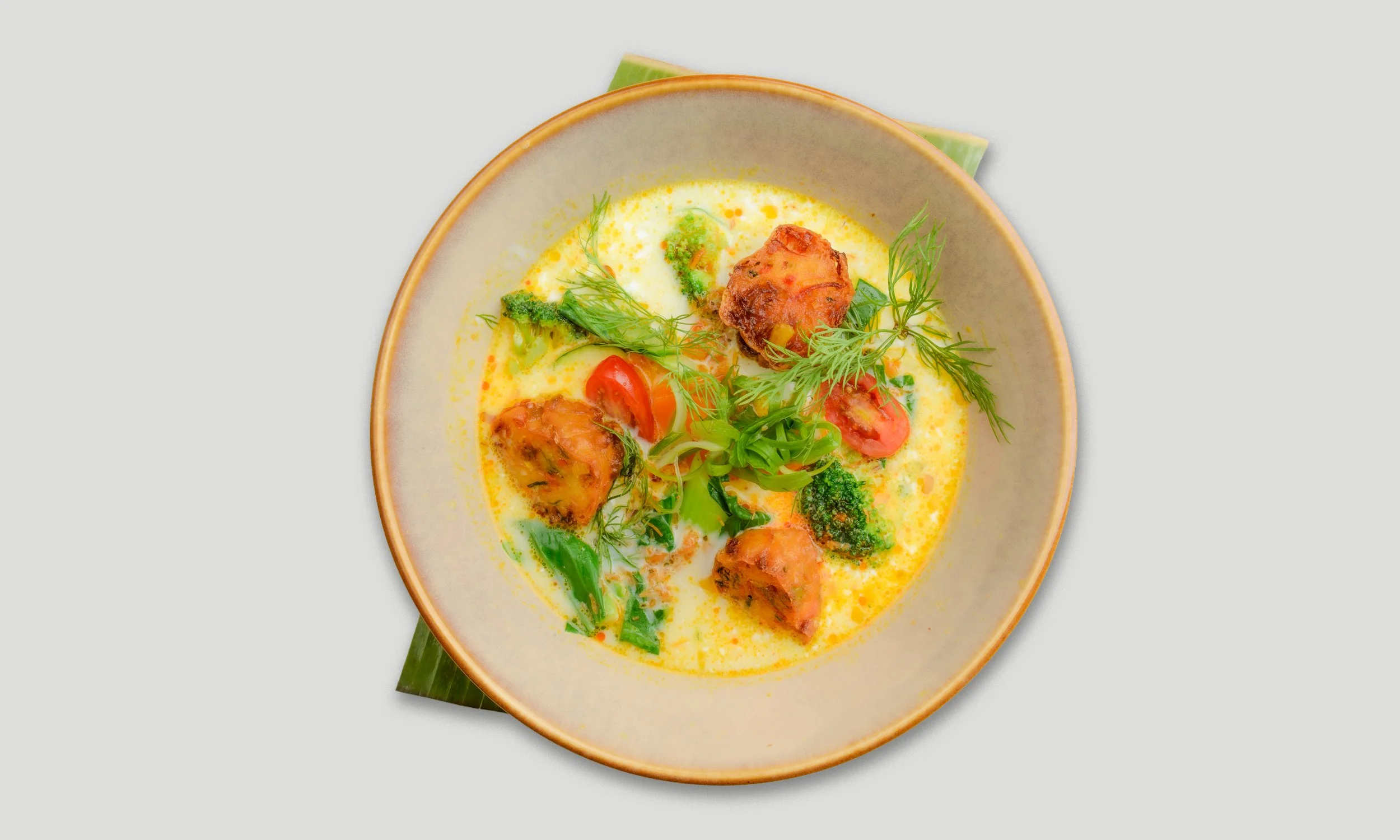Eine Schüssel mit curryartiger Suppe, garnierte mit Frühlingszwiebeln, Tomaten, Brokkoli, und Kräutern, auf einem grünen Blatt.