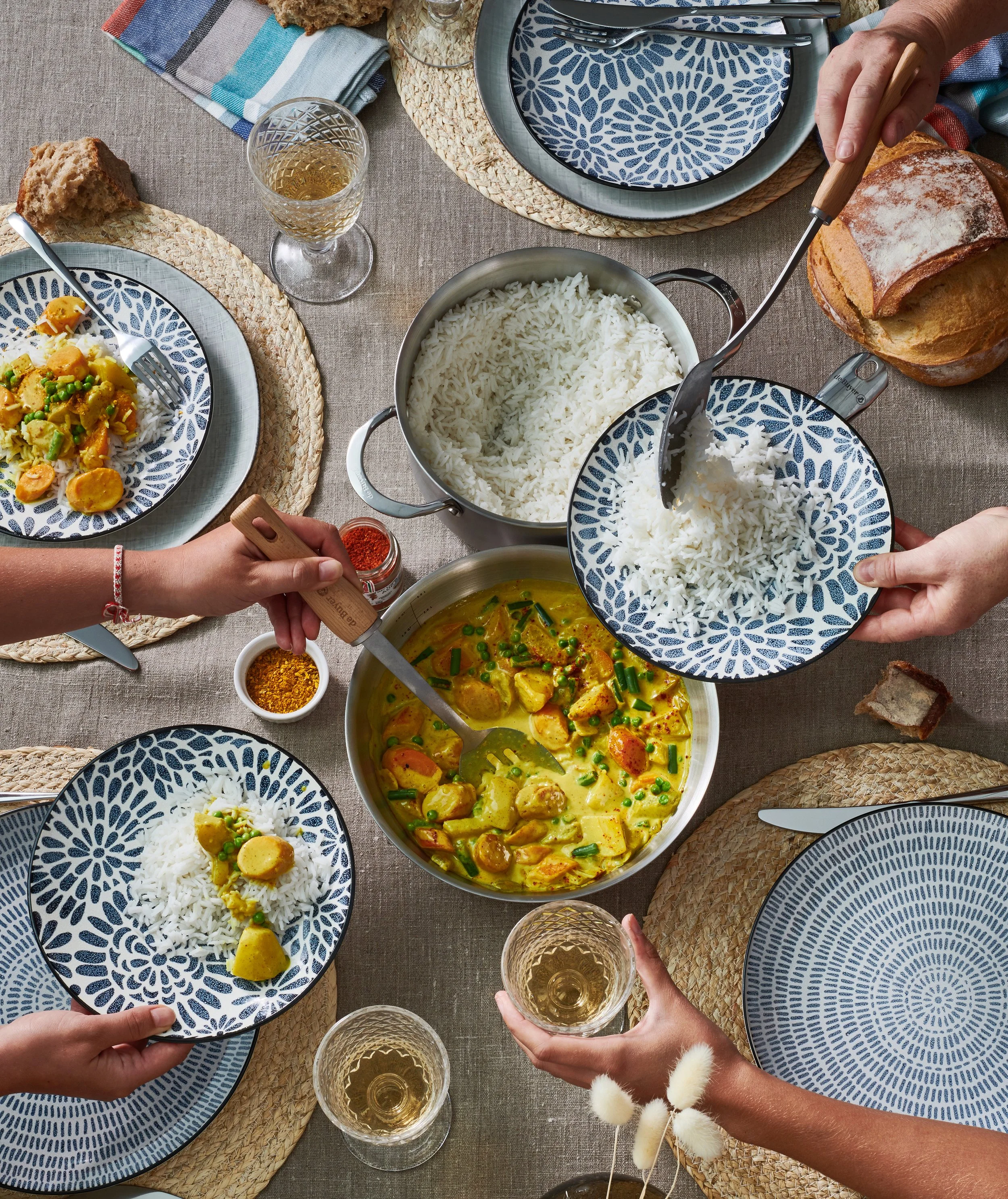 Ein festliches Essen mit weißen Reis, einem Currygericht, frischem Brot und Gläsern Wein. Das Essen ist auf blau-weißen Tellern auf einem großen Holztisch mit Platzdeckchen serviert, und mehrere Hände greifen nach den Speisen und Getränken.