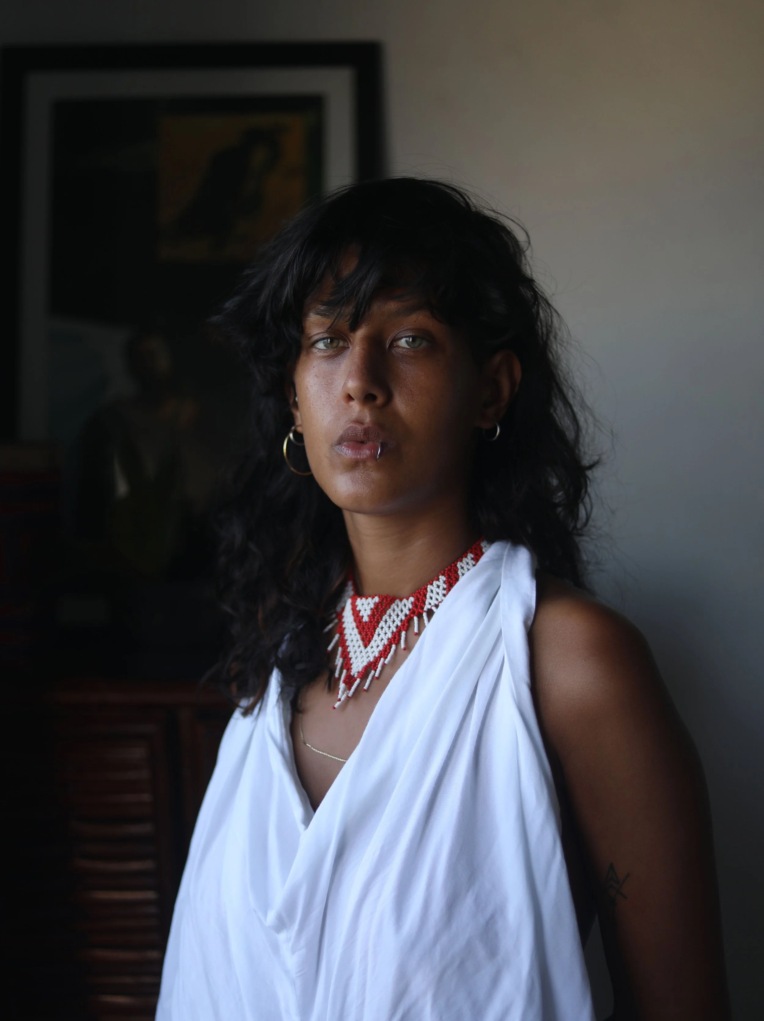 A woman with curly black hair and light skin posing indoors, wearing a white top, beaded necklace, hoop earrings, and a lip piercing.