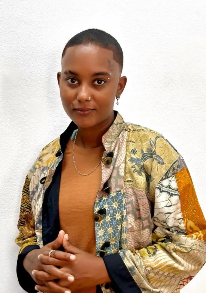 Person with short hair, nose piercing, and eyebrow jewelry, wearing a patchwork patterned jacket over a brown shirt, standing against a white wall.