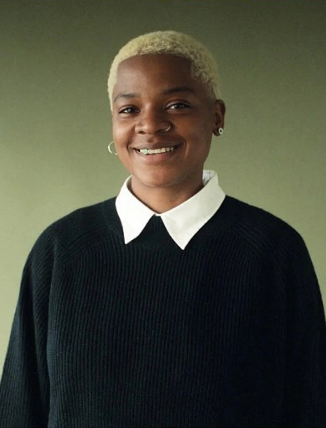 A person with short, blonde hair smiling, wearing a black sweater over a white collared shirt, and standing against a neutral background.