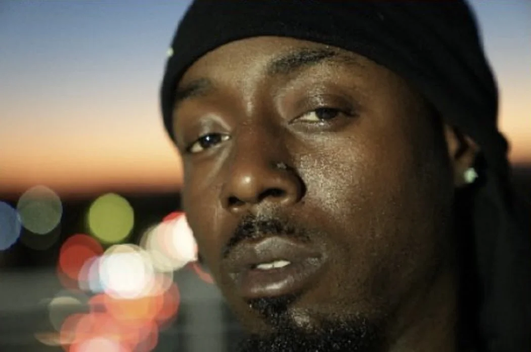 Close-up of a black man wearing a black headscarf, with blurry city lights and sunset in the background.